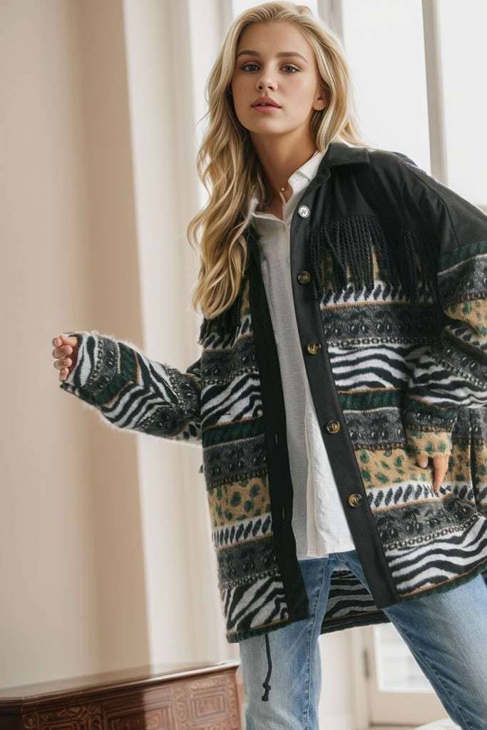 Brown Aztec print shacket with fringe detail, button closure, and patterned sleeves, styled with cream pleated pants while seated on a sofa.