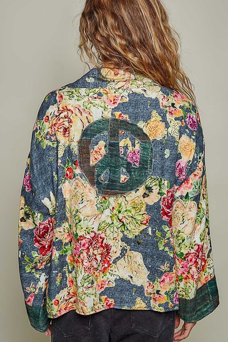 A woman viewed from behind wearing a long-sleeve floral jacket with a large peace sign patch on the back, set against a plain light gray background.