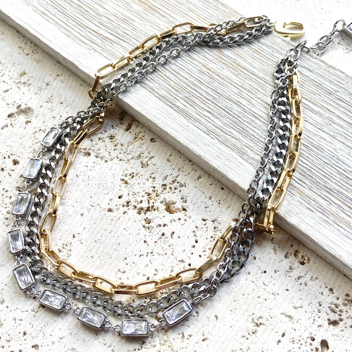 A multi-strand necklace featuring silver and gold-tone chains with rectangular clear crystals, styled on a distressed white wooden board over a textured beige surface.