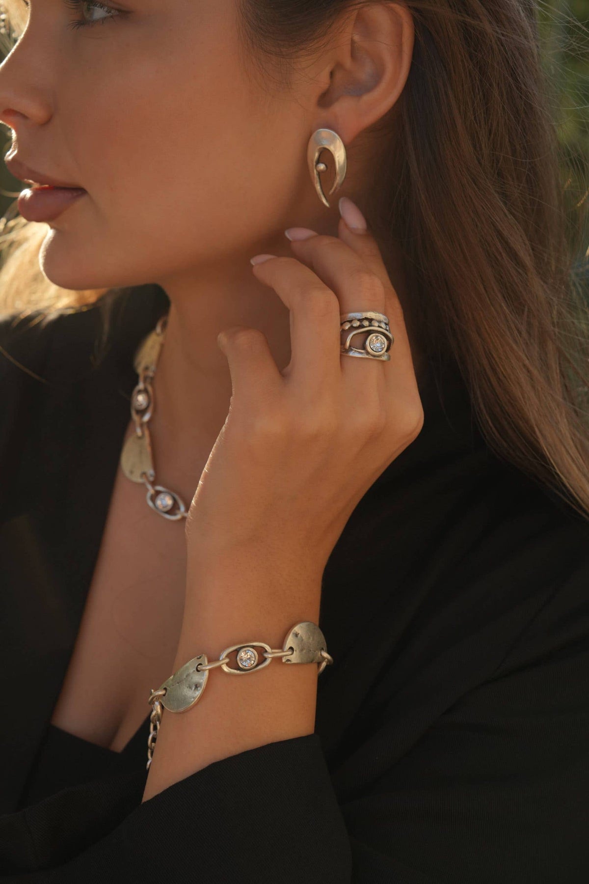 Close-up of a woman wearing gold earrings, bracelet, and ring with a blurred natural background