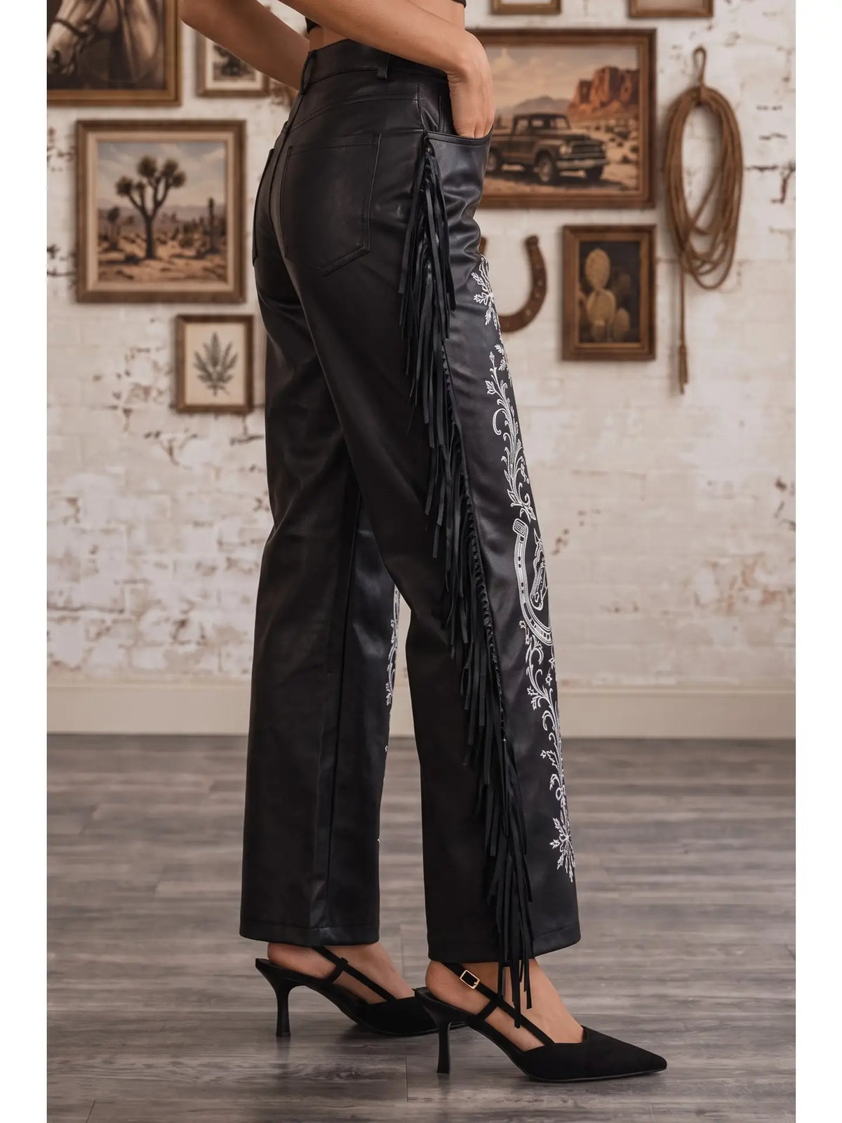 Black faux leather wide-leg pants with long fringe and white floral embroidery down the side, shown on a model wearing black slingback heels in a rustic indoor setting.