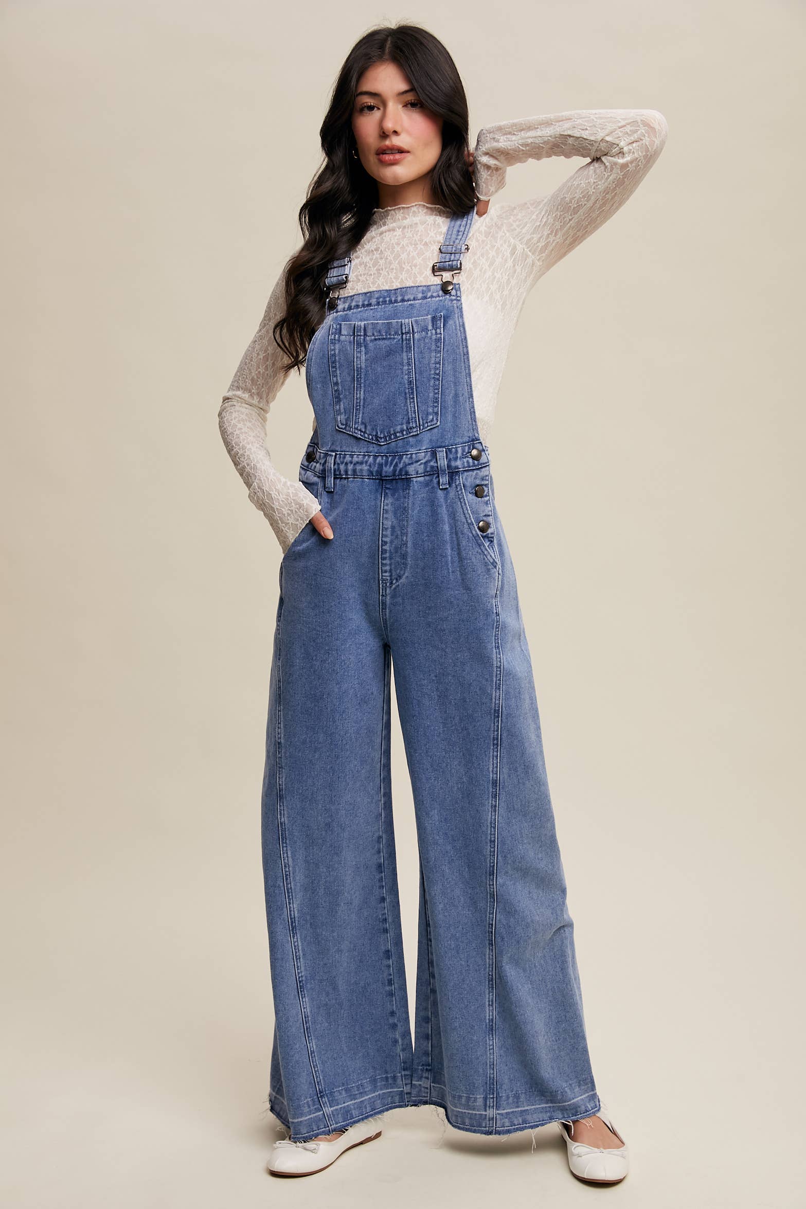 Light-wash wide-leg denim overalls with adjustable straps, front bib pocket, side buttons, and slant pockets, finished with a raw hem and layered over a cream long-sleeve top.