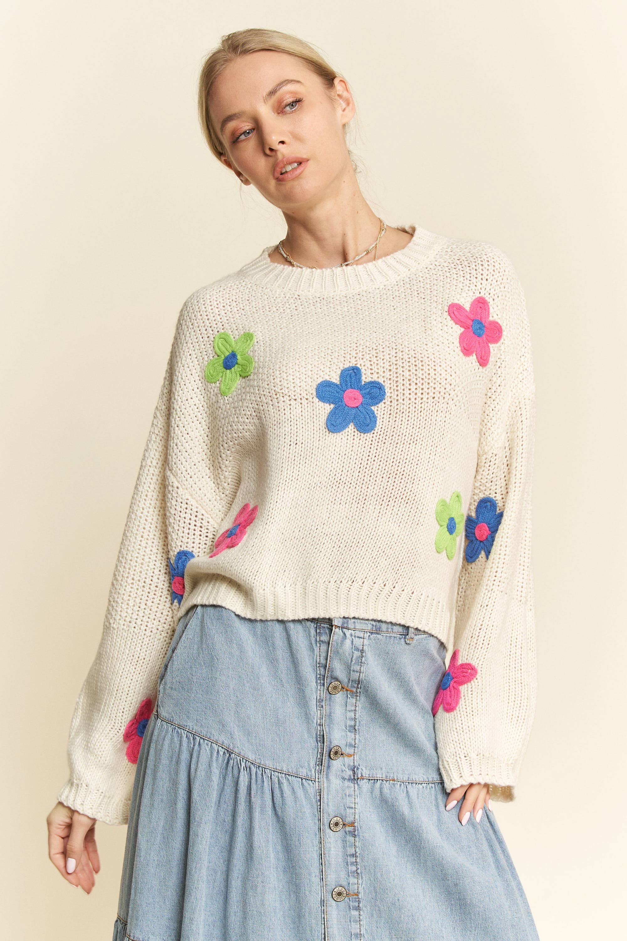 Cream knit sweater with colorful 3D floral appliqués in pink, blue, and green, styled with a button-front light denim skirt.
