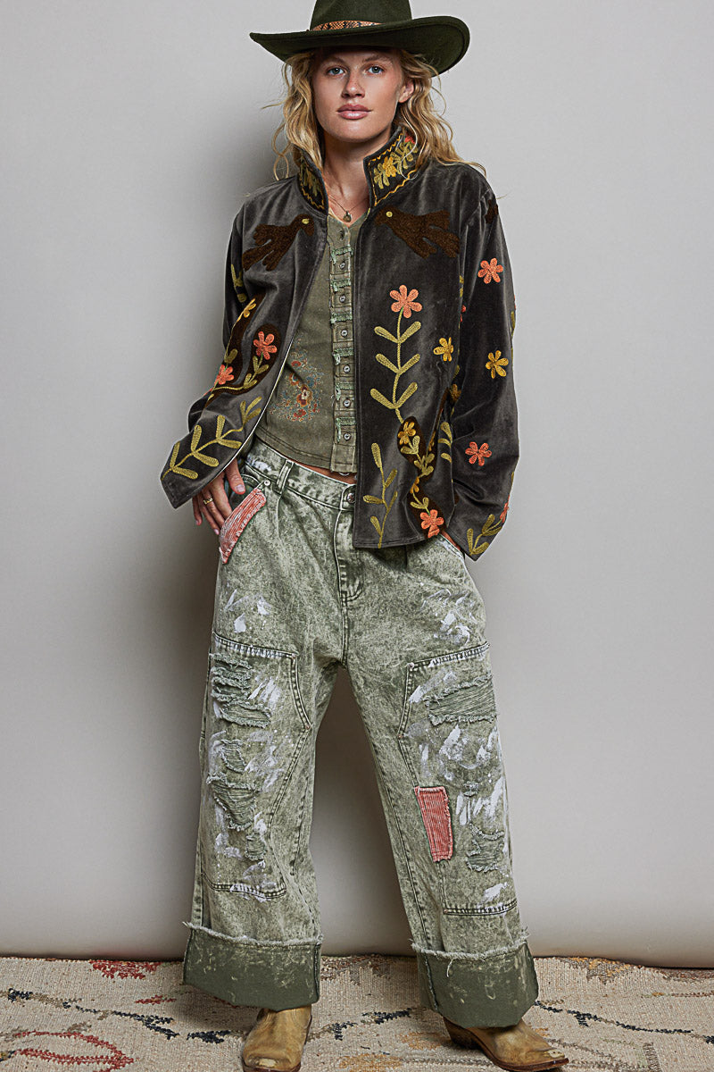 A model wearing a dark brown embroidered velvet jacket, distressed sage green wide-leg pants, a green hat, and brown boots, standing against a neutral gray wall.