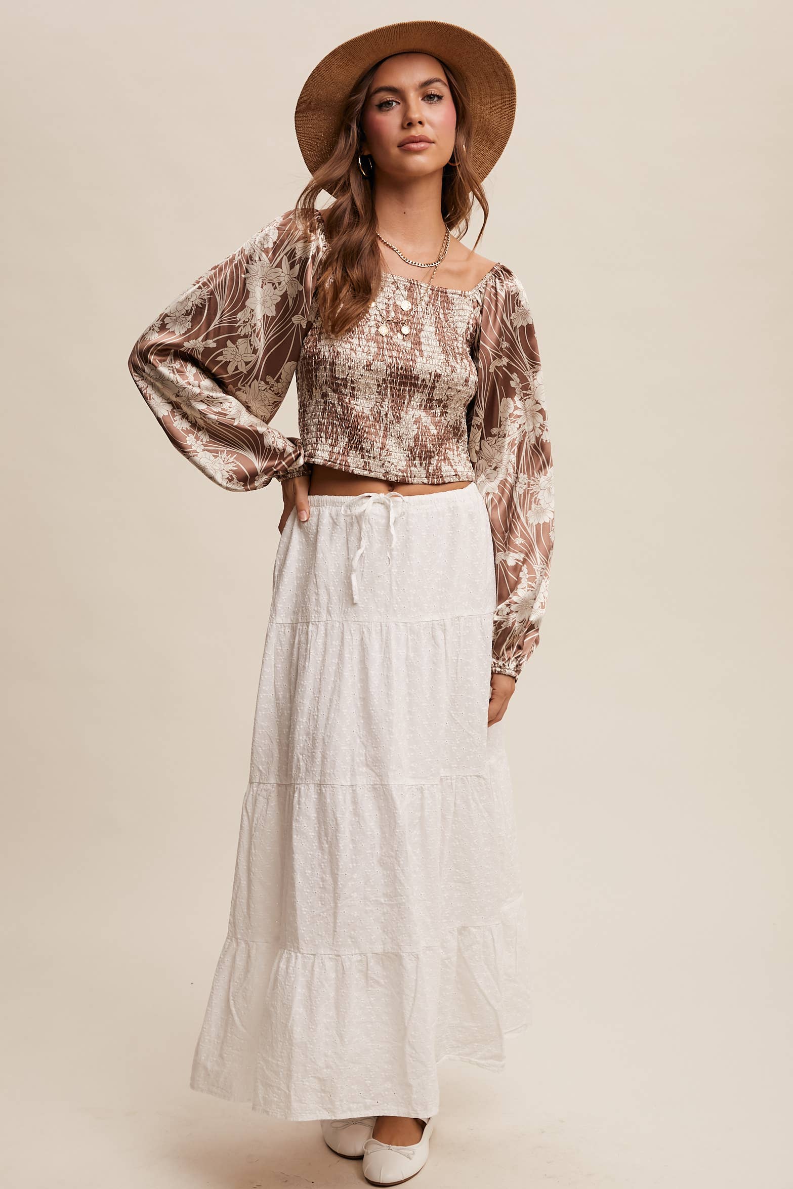 Brown and cream floral smocked blouse with square neckline, puffed long sleeves, and elastic cuffs, styled with layered necklaces and a wide-brim hat.