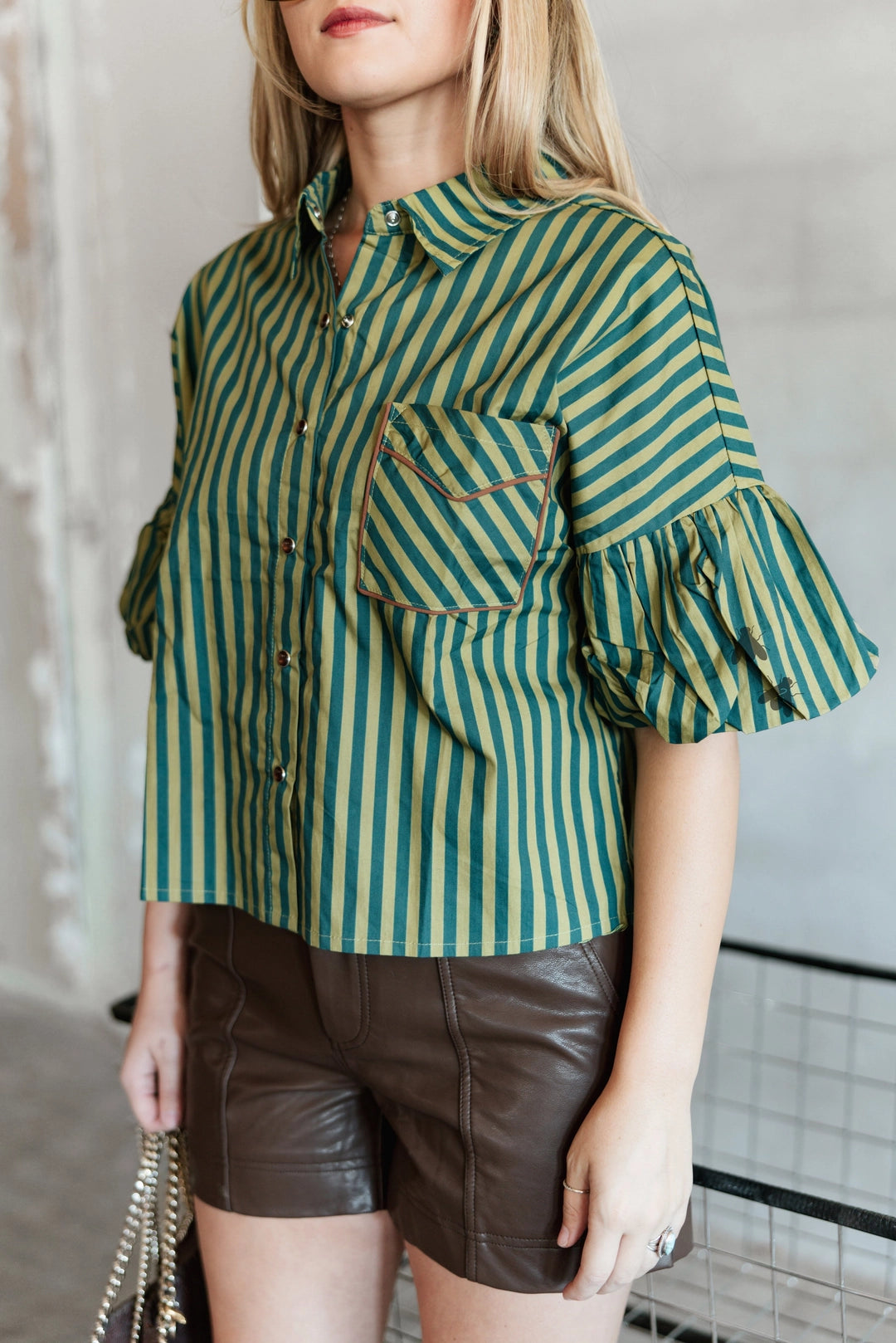A woman wearing a green and teal striped short-sleeve button-up shirt with ruffled sleeves and brown faux-leather shorts, holding a gold chain handbag.