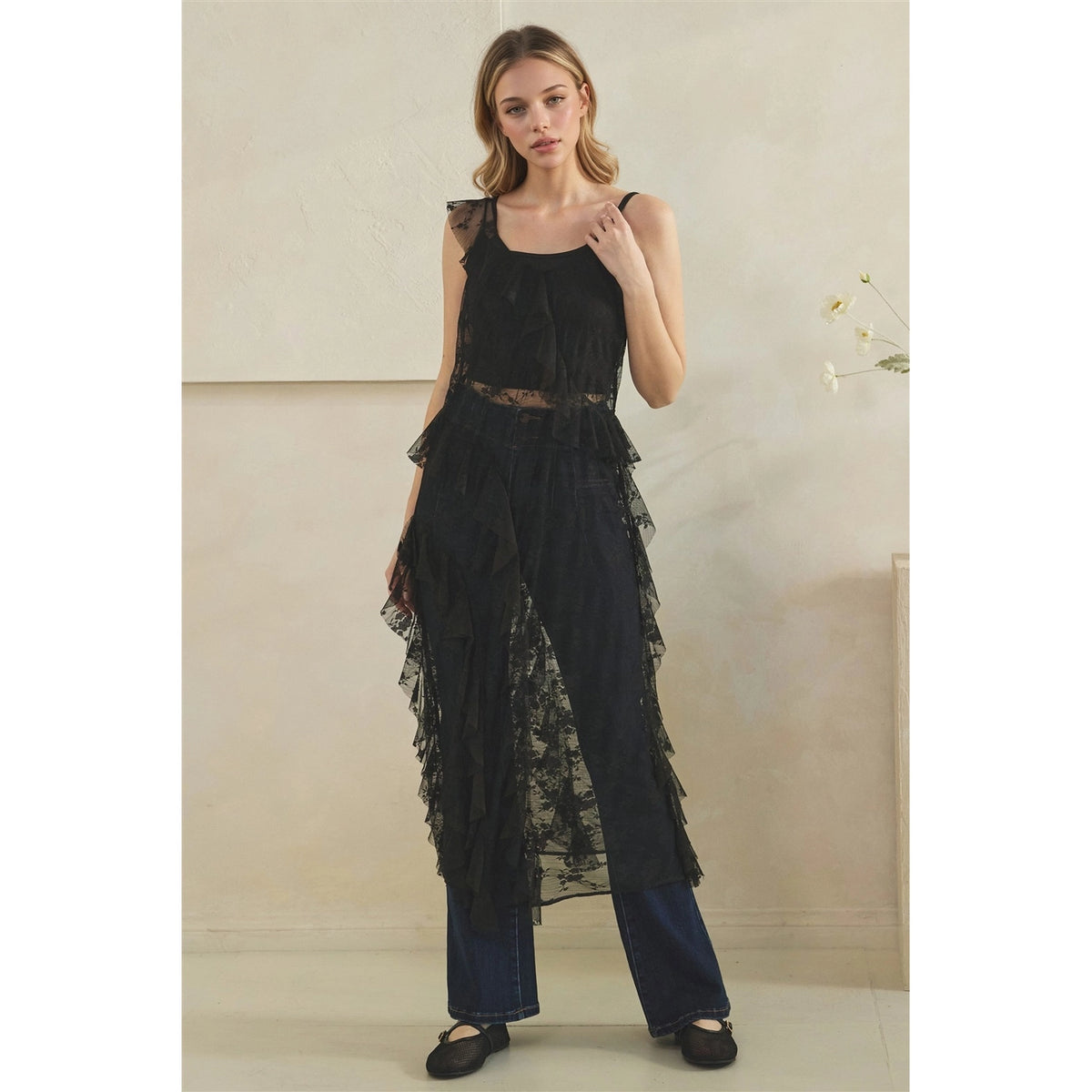 A woman models a black sheer lace sleeveless long ruffled duster over dark black wide-leg pants, standing against a neutral beige wall.