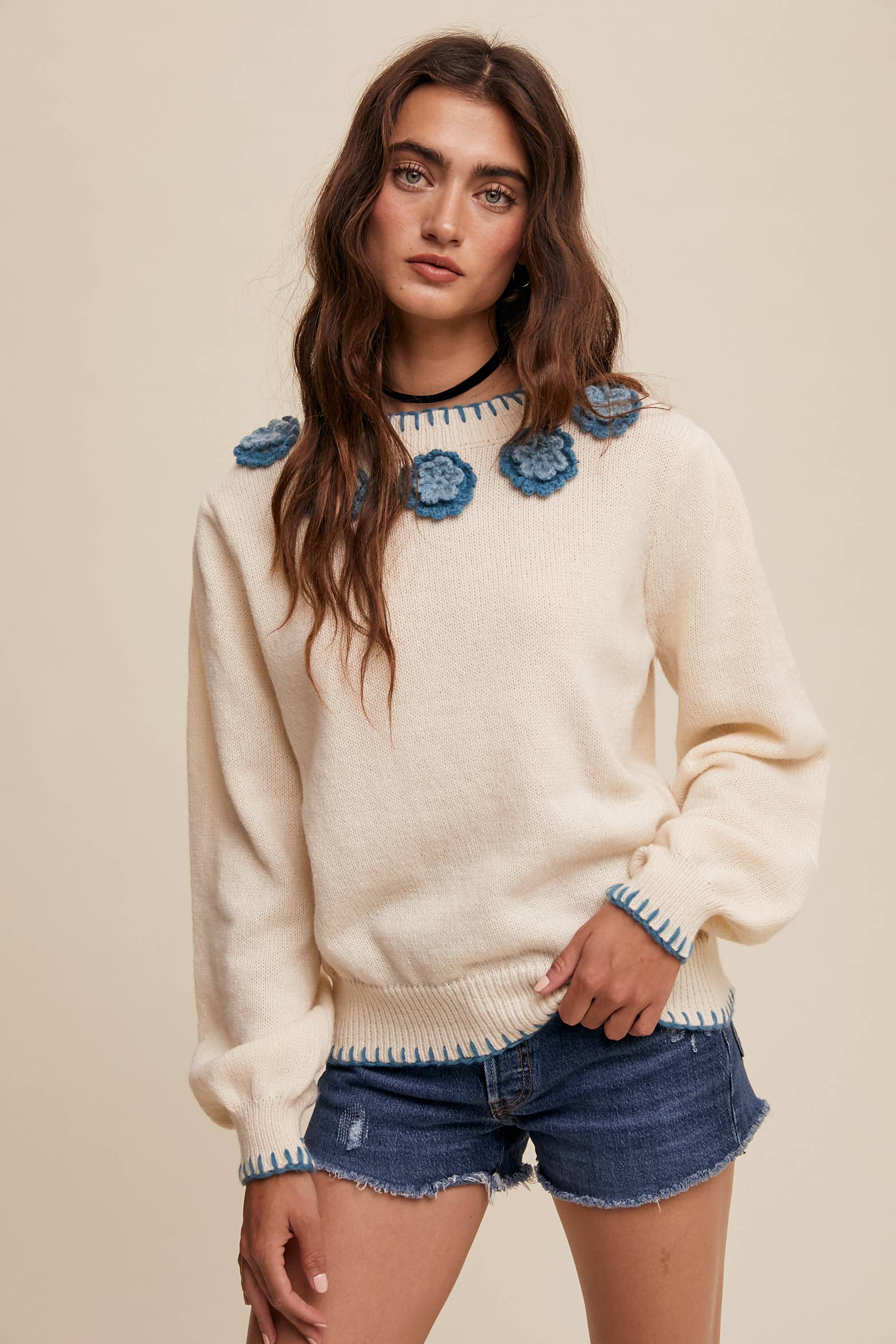 Cream sweater with blue crochet flower appliqués at neckline, blue whipstitch trim at cuffs and hem, long sleeves; styled with frayed denim shorts.