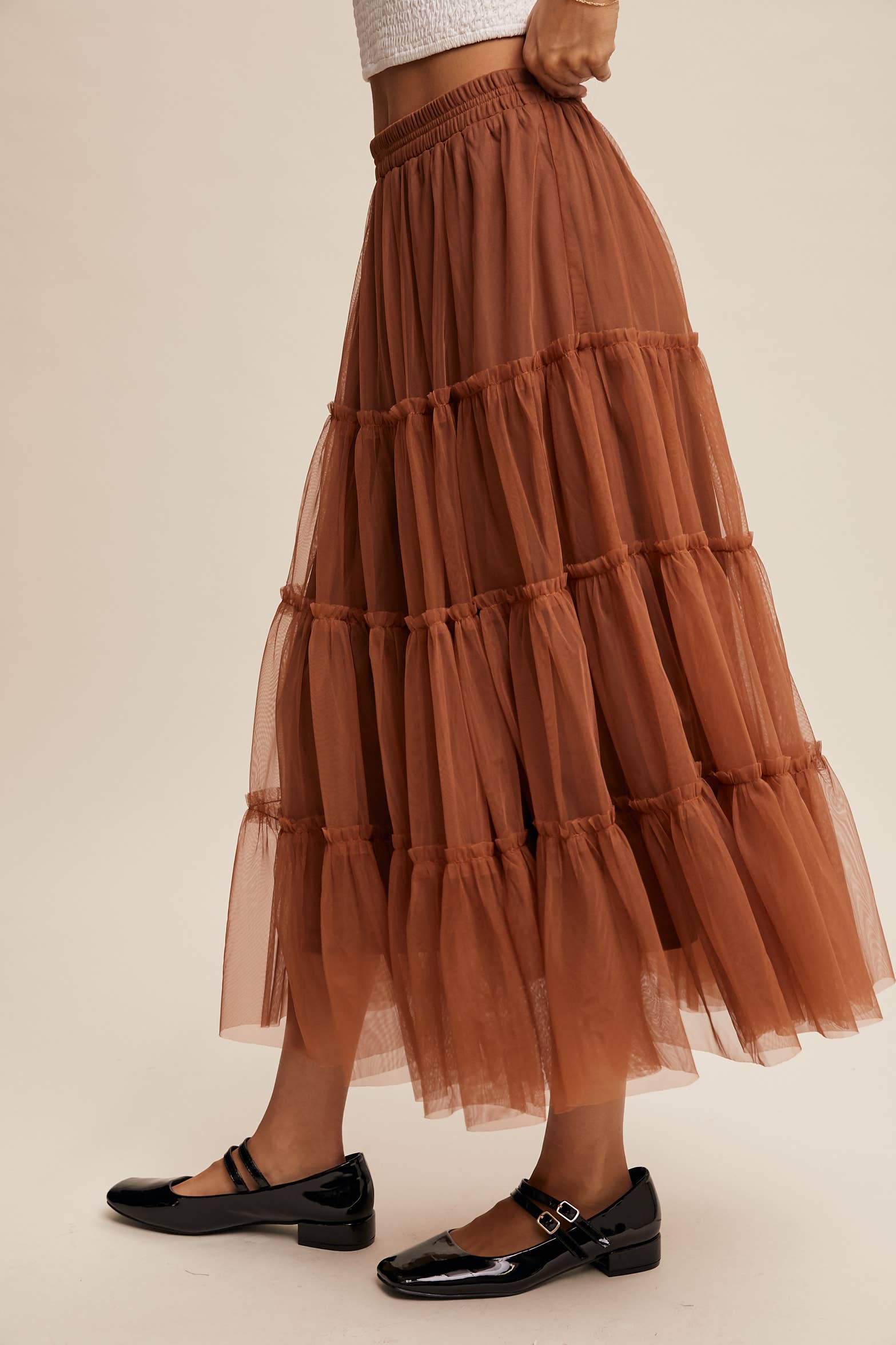 Side view — rust tulle tiered midi skirt with elastic waist and ruffle tiers, sheer overlay; model in black Mary Jane flats.