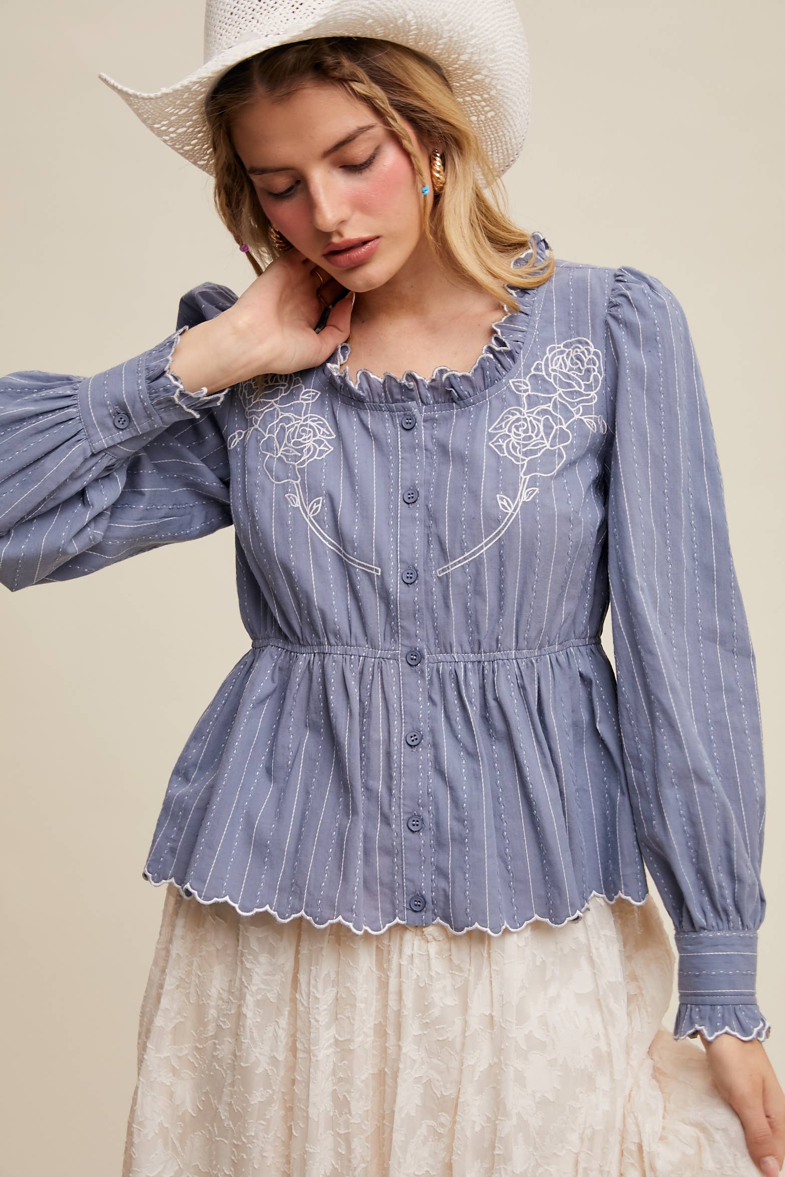 Dusty blue pinstripe blouse with ruffled neckline, white rose embroidery, puff long sleeves, button front, and scalloped peplum hem—styled with a white lace skirt and cowboy hat.