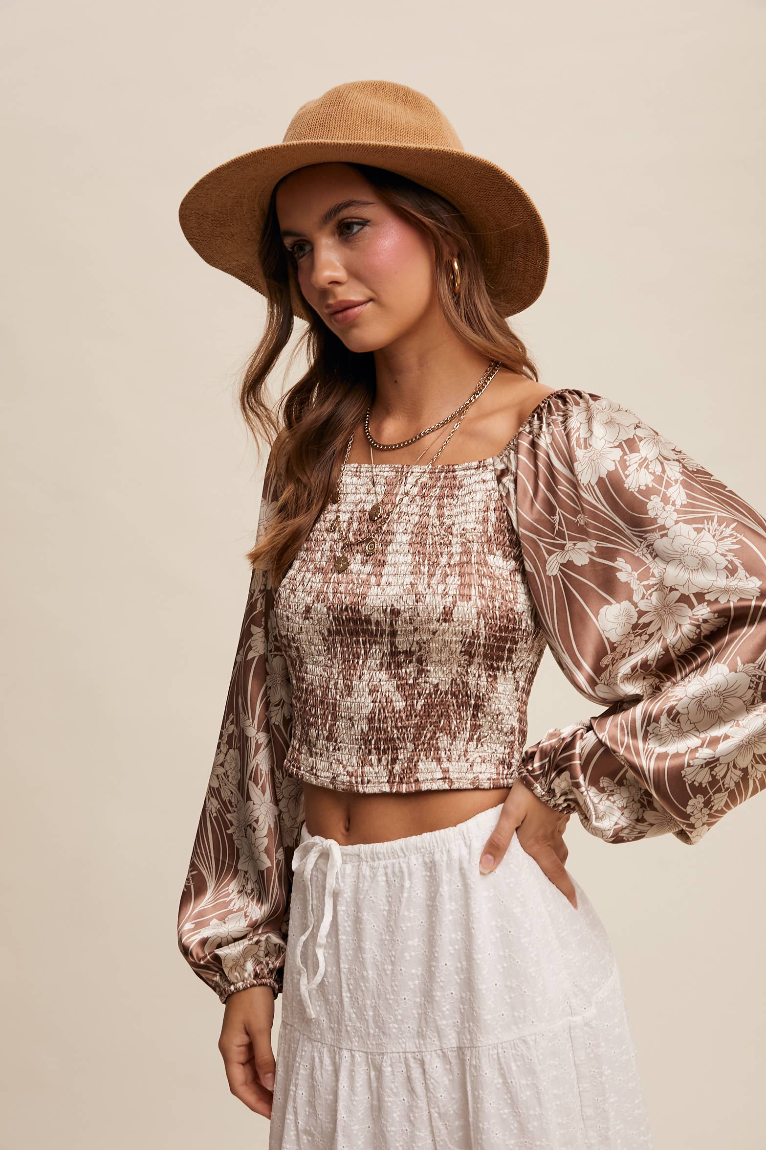 Brown and cream floral smocked blouse with square neckline, puffed long sleeves, and elastic cuffs, styled with layered necklaces and a wide-brim hat.