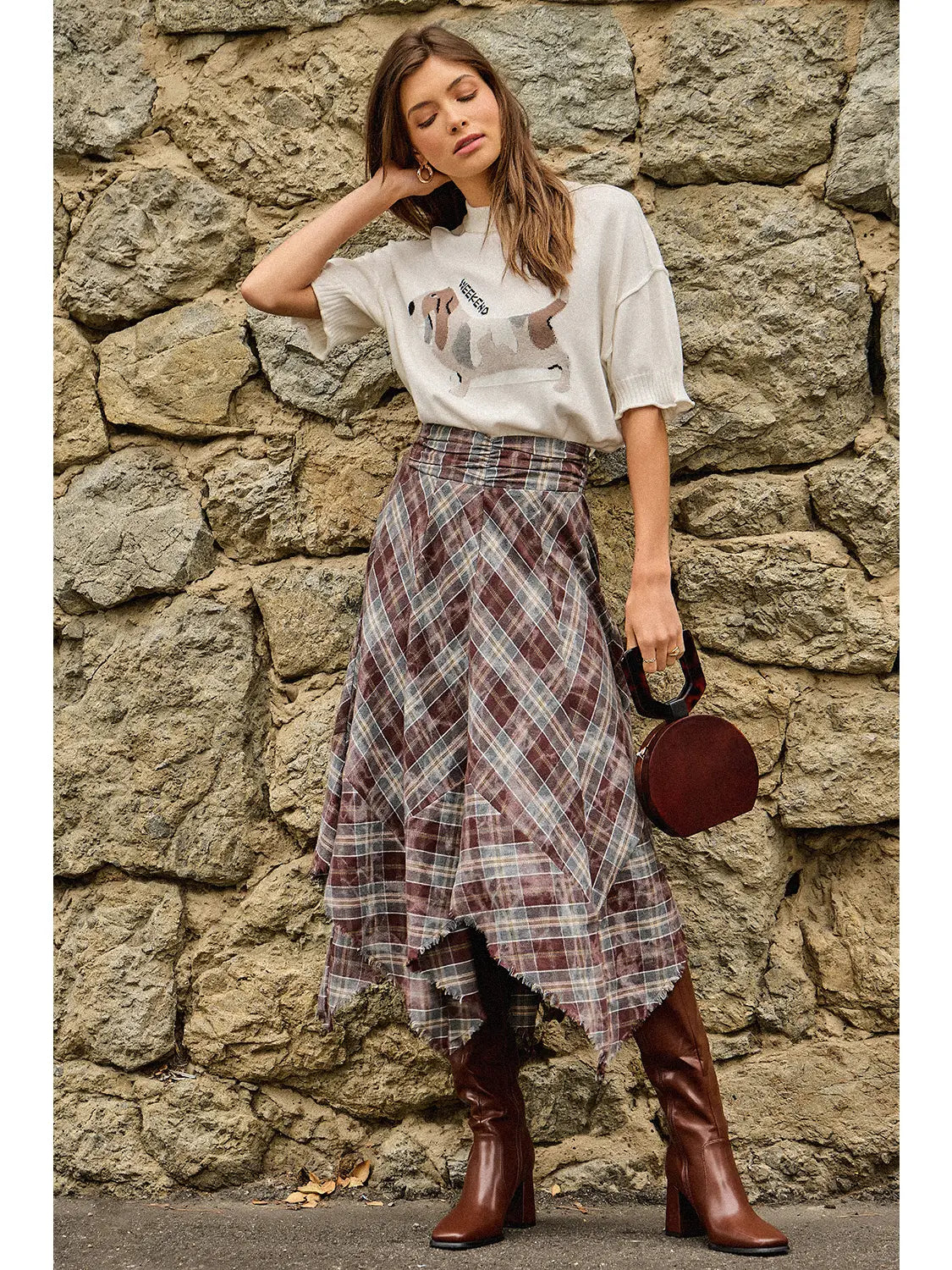 A woman stands against a rustic stone wall wearing a white graphic T-shirt, a brown and gray plaid wrap skirt, and brown knee-high boots, while holding a small dark red handbag.