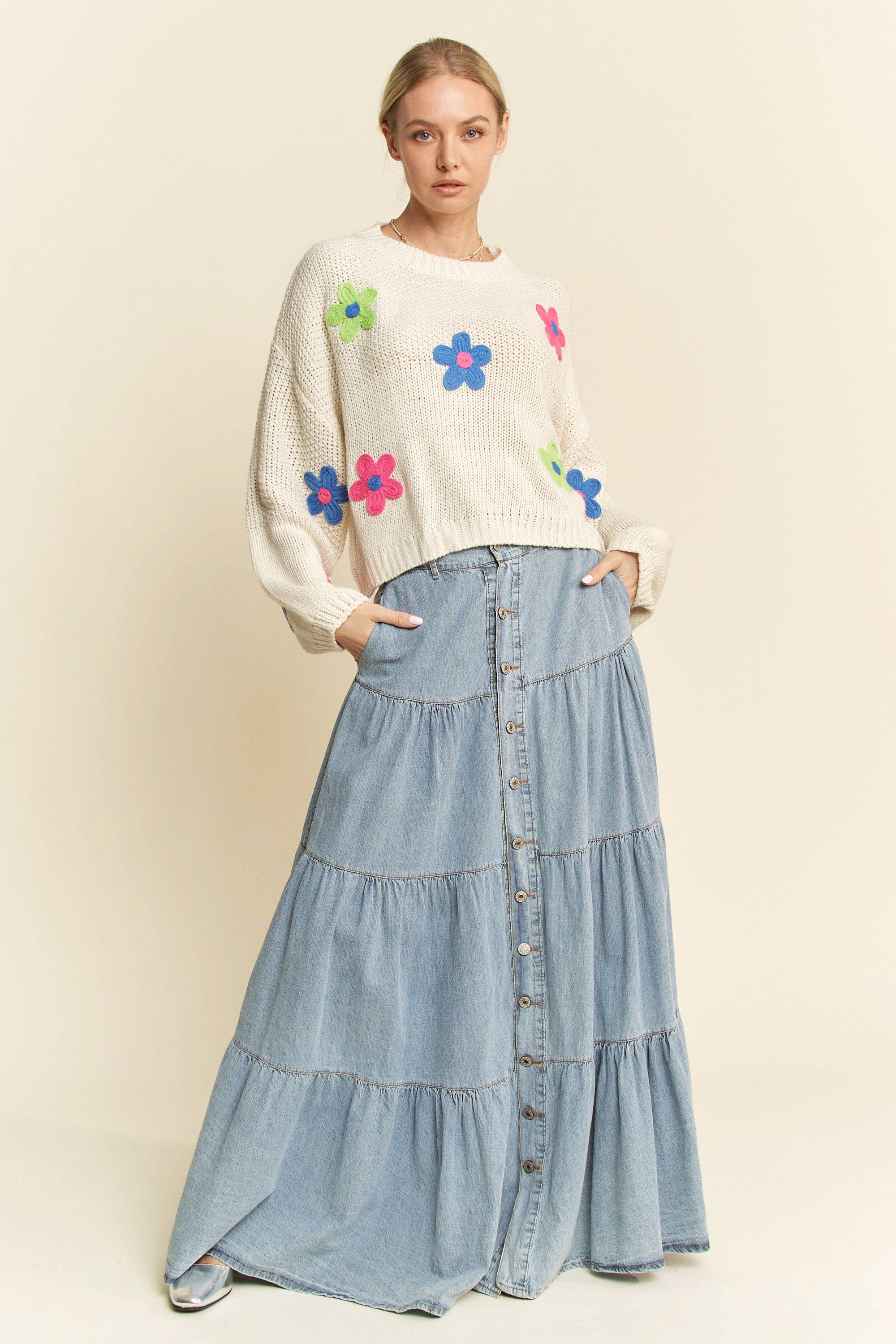 Cream knit sweater with colorful 3D floral appliqués in pink, blue, and green, styled with a button-front light denim skirt.