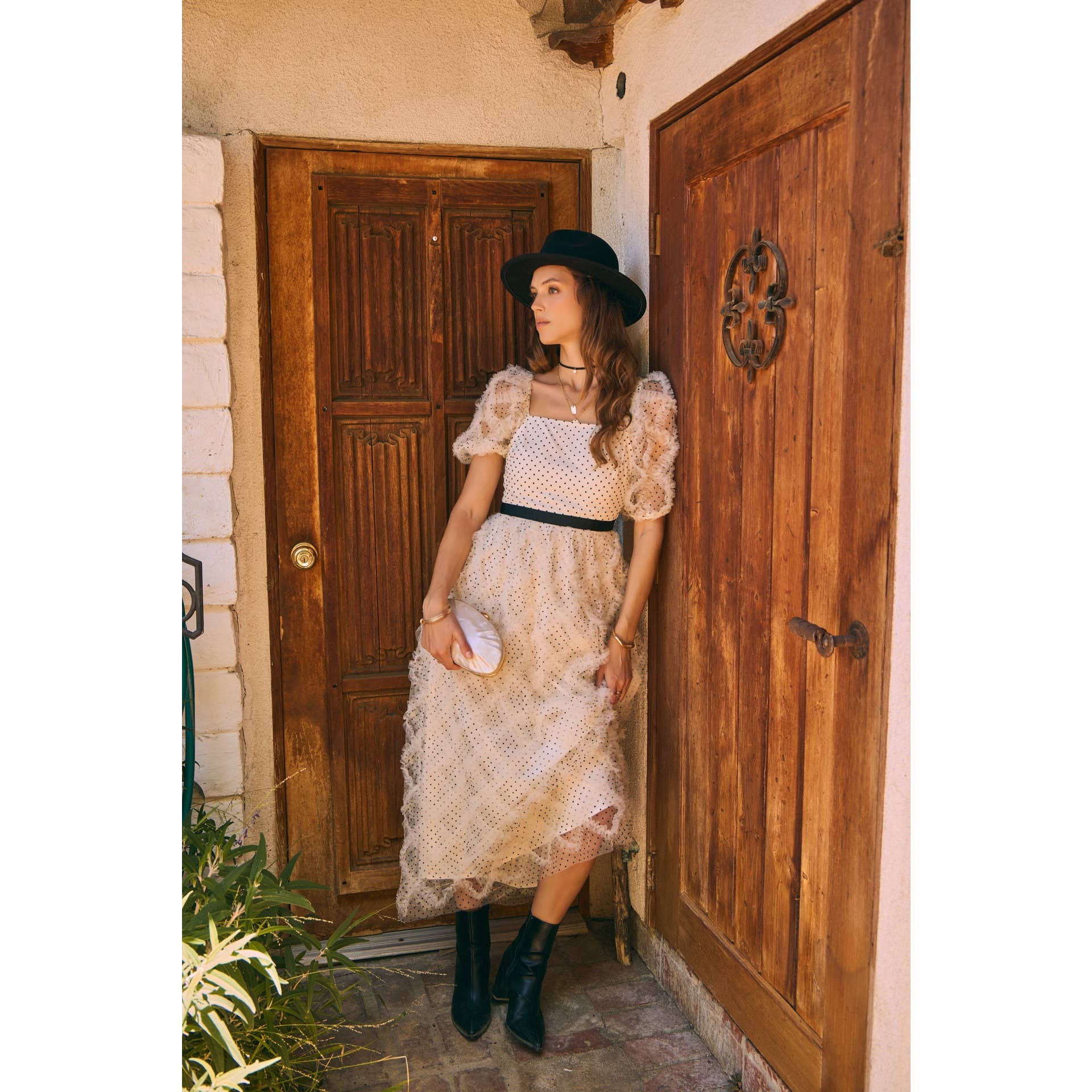 Front view — cream polka-dot tulle midi dress with puff sleeves and black belt; styled with black hat, ankle boots, pearl clutch; leaning by wooden doors.