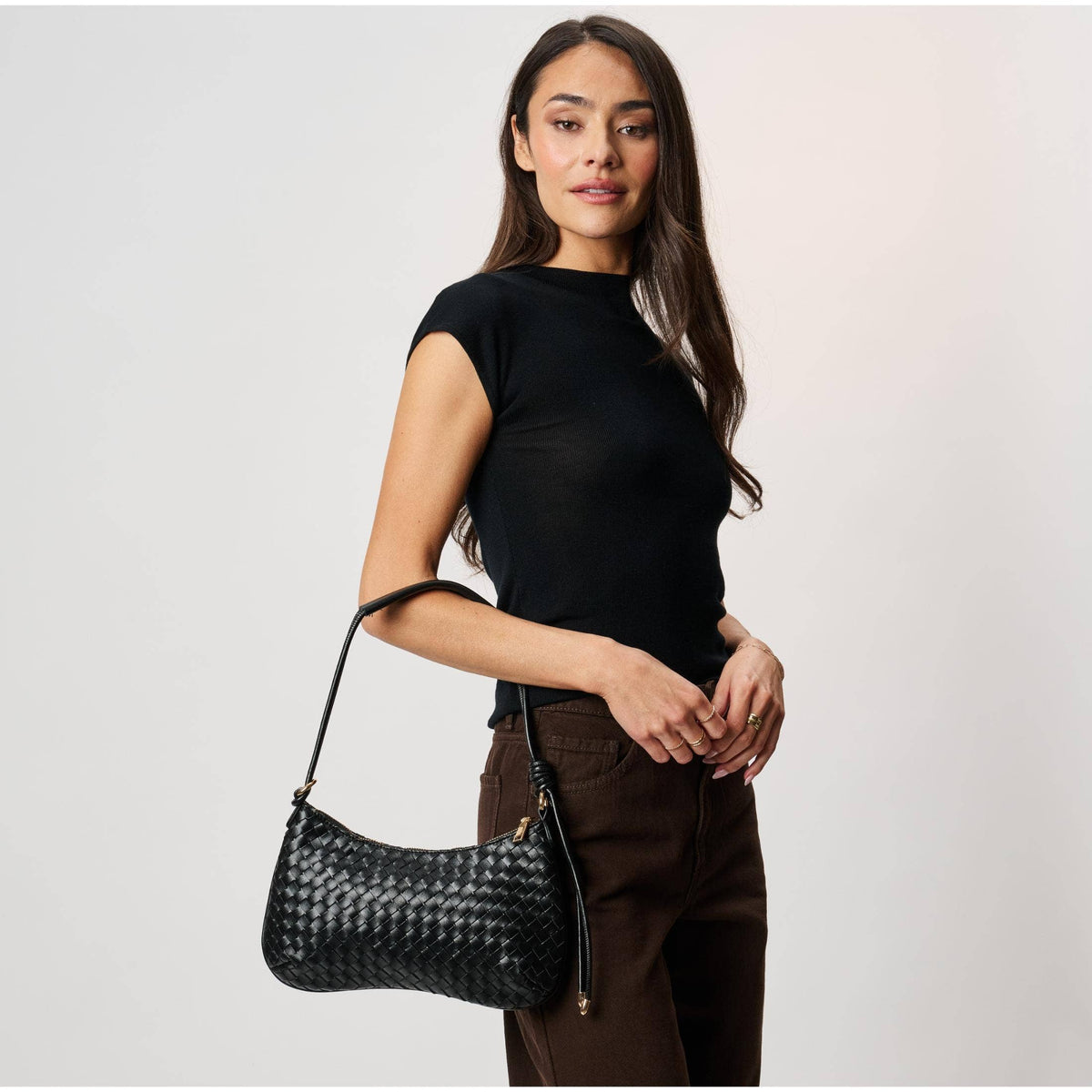 Cindy Woven Crescent Shoulder Bag in Black by Moda Luxe — close-up model carry view showing woven vegan leather crescent silhouette, knotted strap detail, and gold-tone zip pull, styled with black top and denim jeans