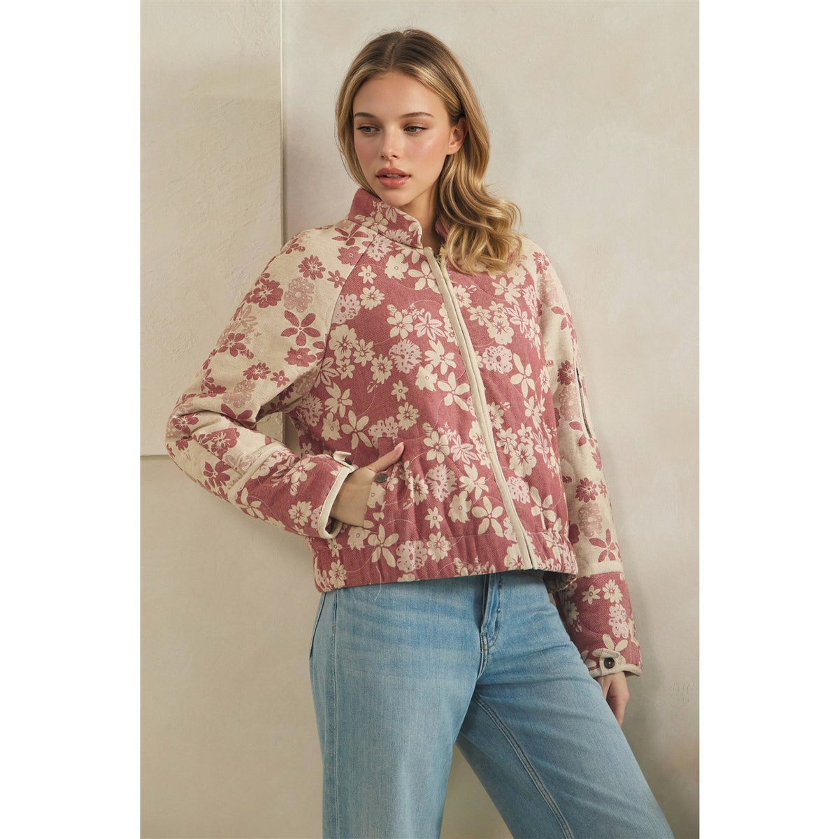 A woman models a cropped zip-front jacket with a dusty rose and cream floral pattern, paired with light blue jeans, set against a neutral beige wall.