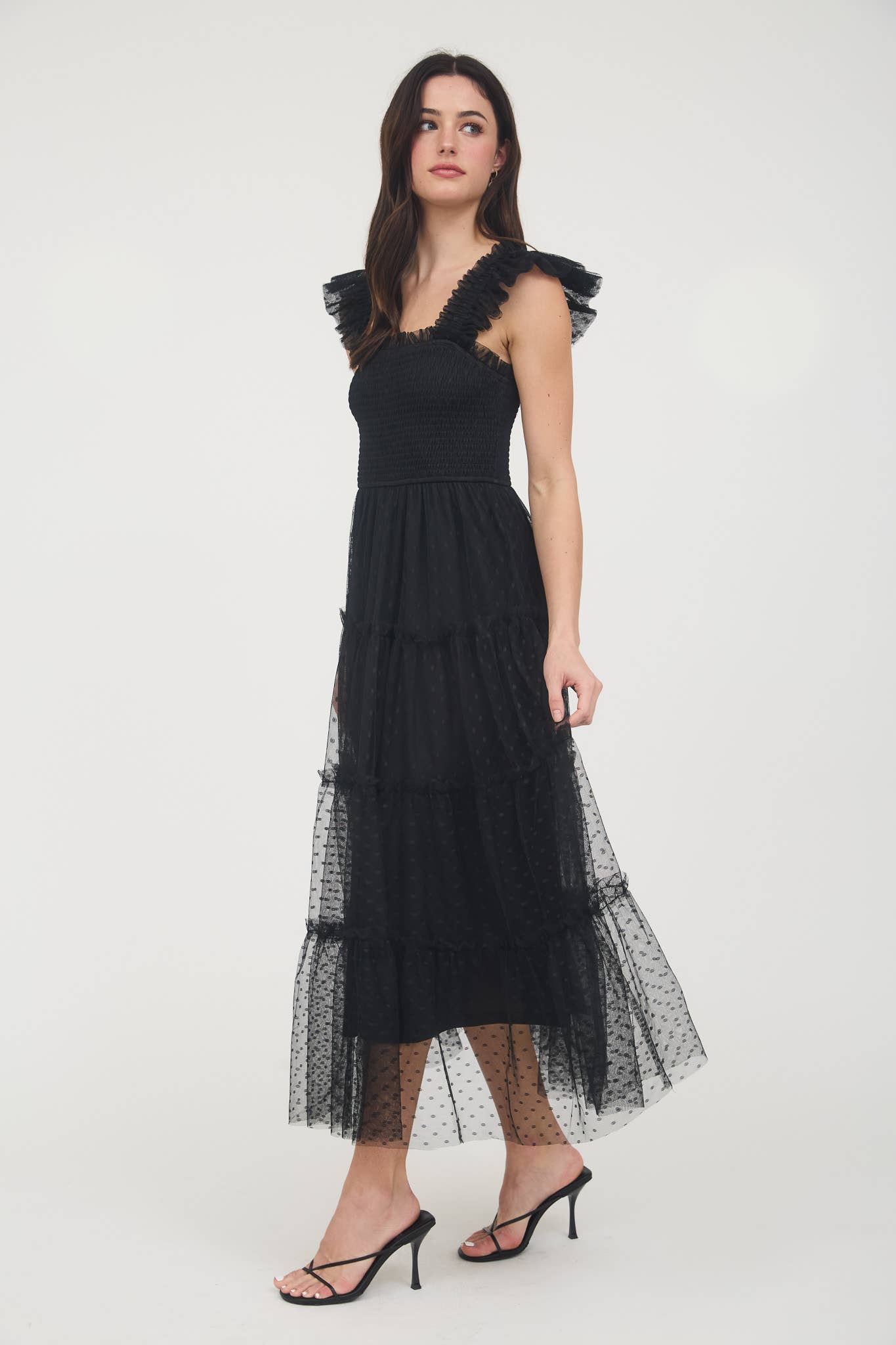 Side view of Model wearing black
 smocked midi dress with ruffle sleeves, tiered dotted mesh overlay, and flowy A-line silhouette.