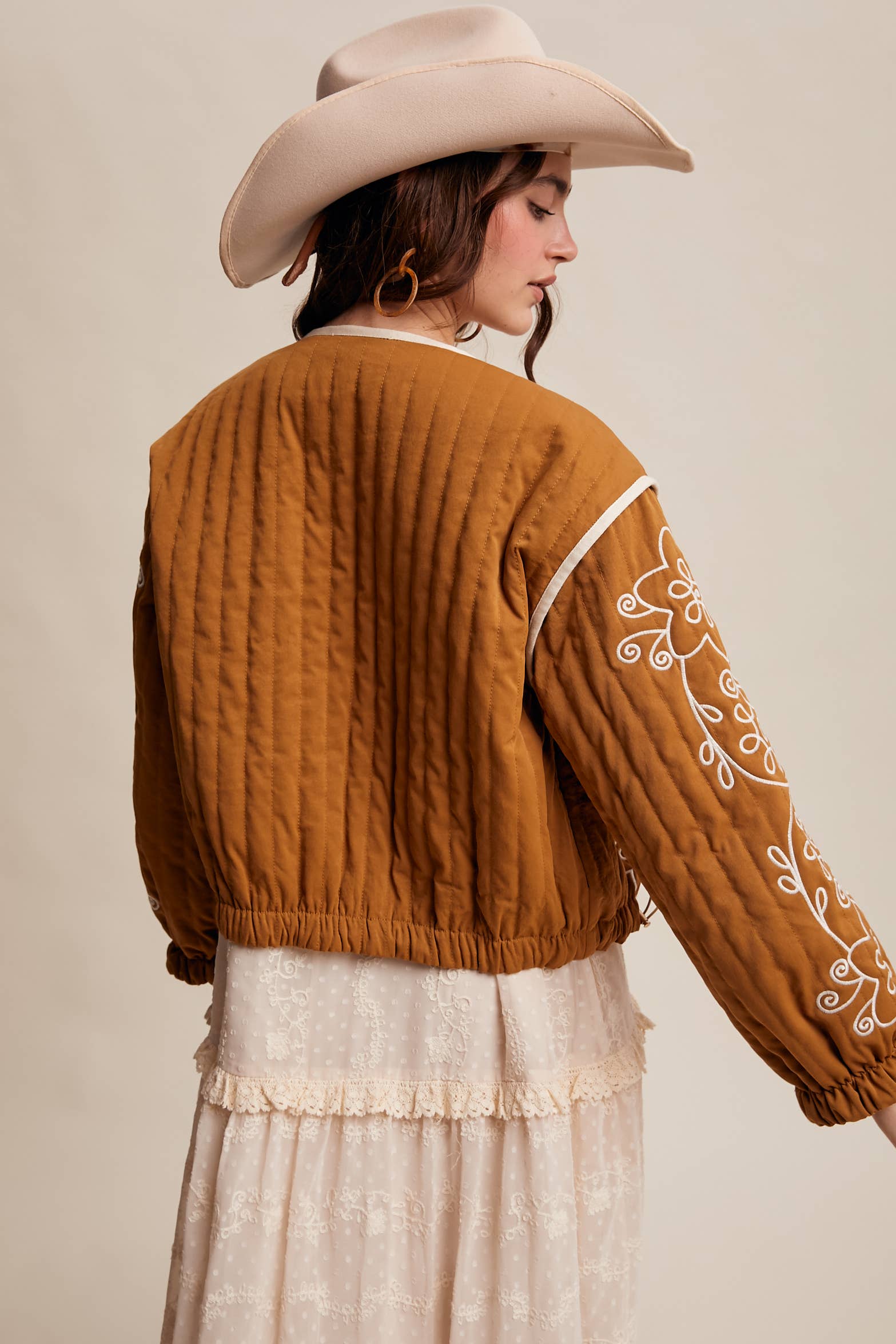 Back view- Boho caramel-brown jacket with cream floral swirl embroidery, ivory piping, and snap buttons, styled over a lace tie-front top with a scarf and felt hat outdoors.