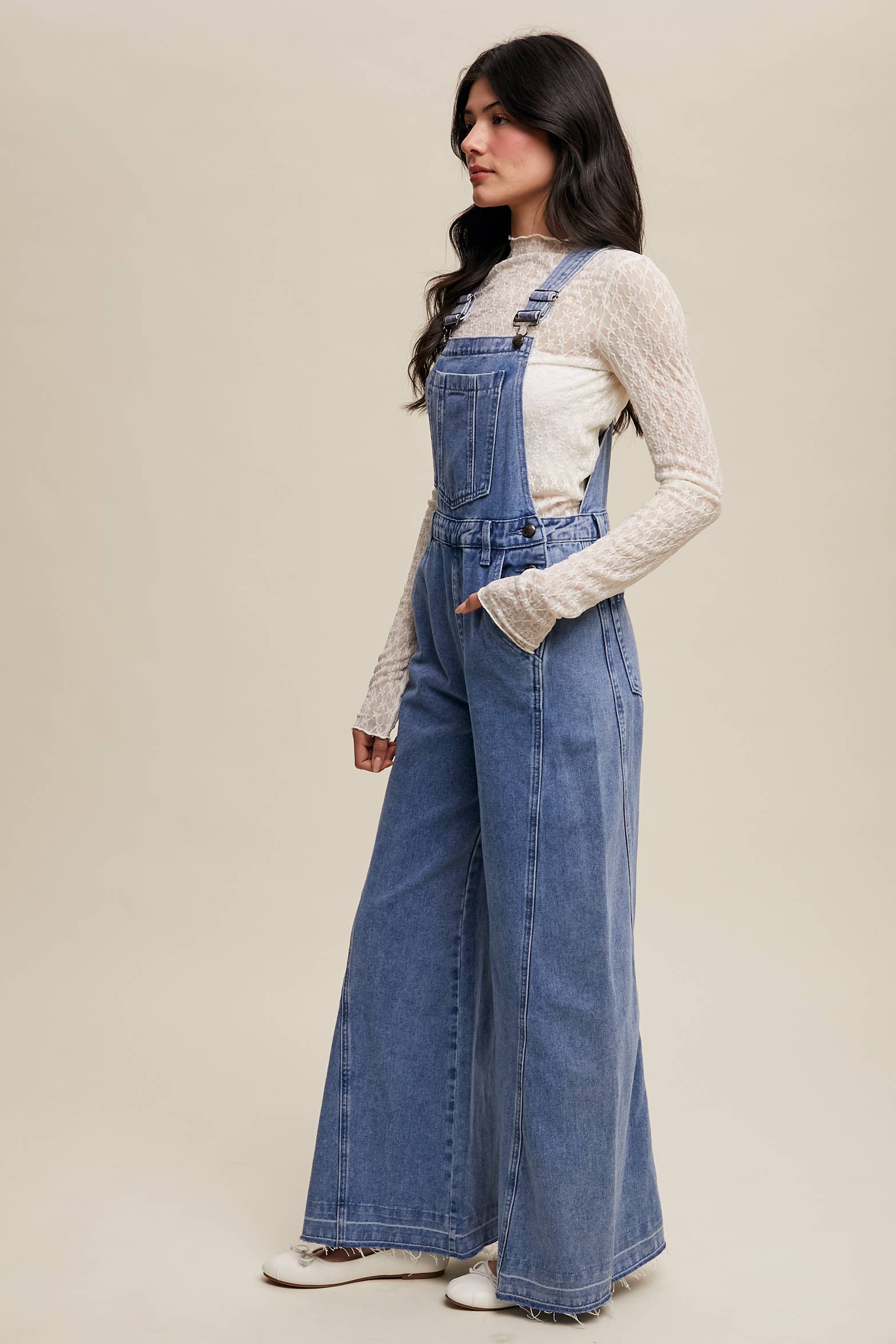 Side view- Light-wash wide-leg denim overalls with adjustable straps, front bib pocket, side buttons, and slant pockets, finished with a raw hem and layered over a cream long-sleeve top.