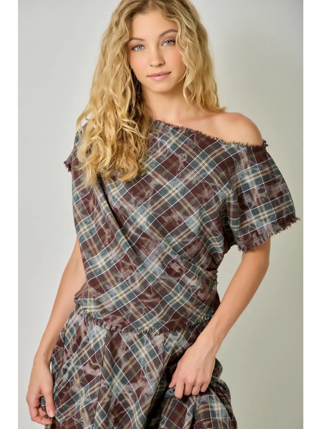 A woman modeling an off-the-shoulder wrap dress with a brown, beige, and blue plaid pattern against a neutral gray background.