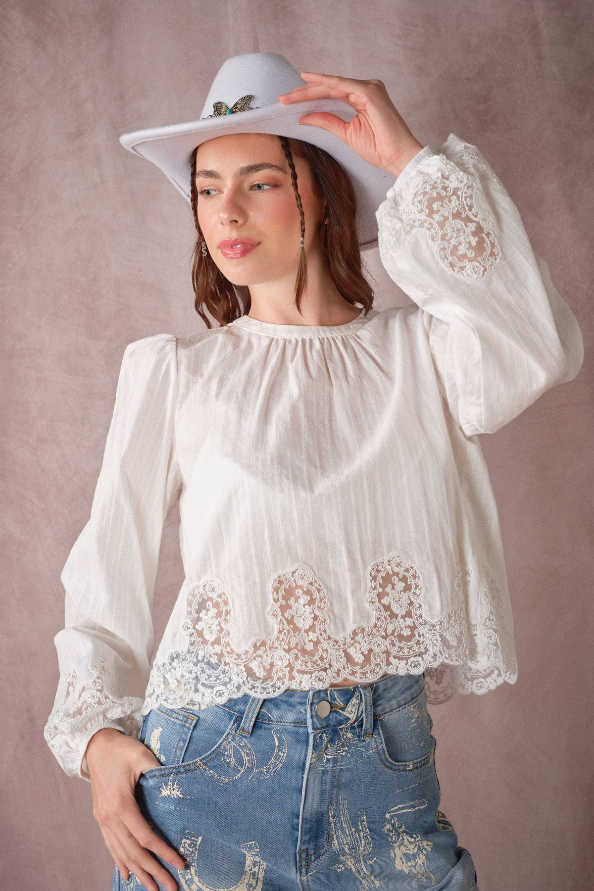 A woman wearing a white long-sleeve blouse with intricate lace detailing at the cuffs and hem, paired with distressed blue jeans and a white wide-brim hat, posing against a textured mauve background.