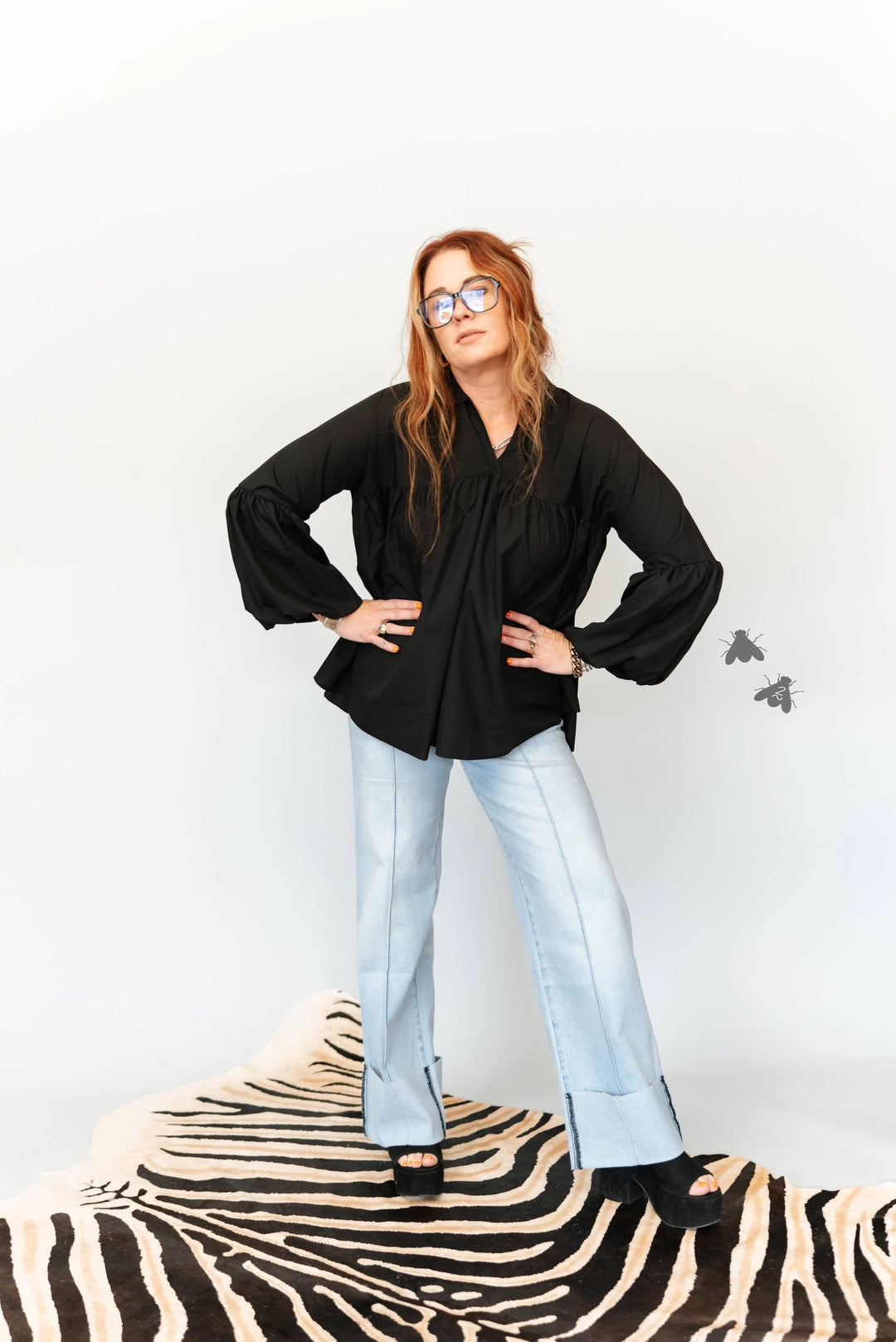 A woman with red hair and glasses wearing a black long-sleeve blouse and light blue jeans, standing on a black and white zebra-patterned rug against a white studio background.