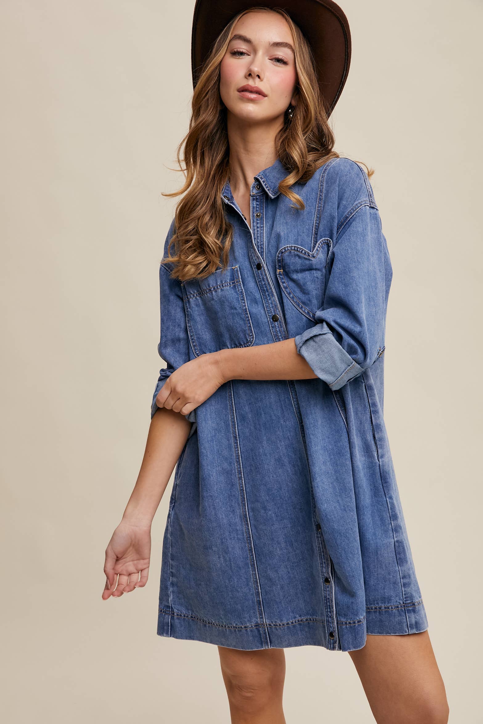 Medium-wash denim shirt dress with snap button front, heart-shaped chest pocket, rolled long sleeves, and a relaxed mini silhouette.