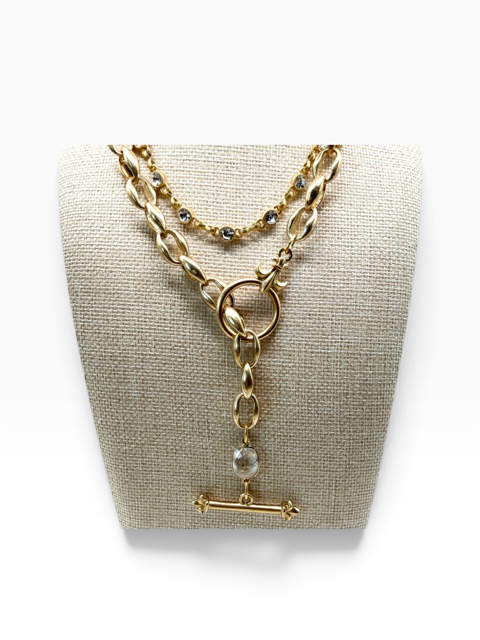 A gold-tone layered necklace displayed on a textured beige fabric jewelry bust. The necklace features a thick twisted chain, a thinner chain with small dark stones, and a Y-shaped drop with a round bead and a horizontal bar pendant.