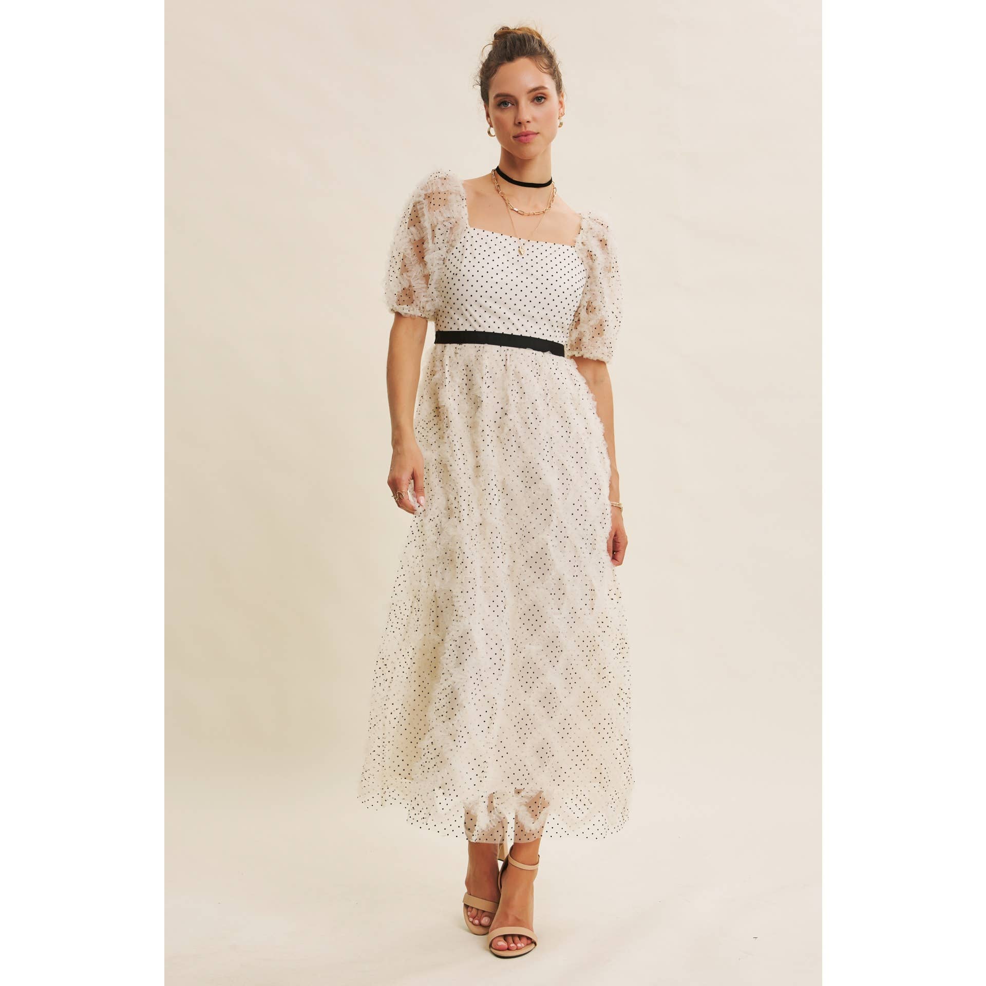Front view — cream polka-dot tulle midi dress with puff sleeves and black belt; styled with black hat, ankle boots, pearl clutch; leaning by wooden doors.