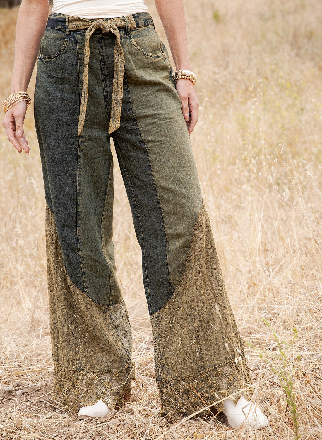 A person standing in a dry grass field wearing wide-leg patchwork pants with dark green denim and tan lace-like lower panels, along with a cream top and white shoes.