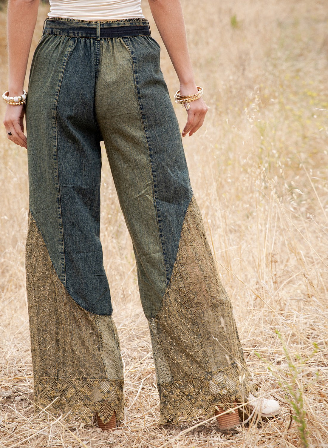 Patchwork wide-leg pants with a dark blue denim upper section and a sheer beige lace lower section, shown on a model standing in a dry grass field.