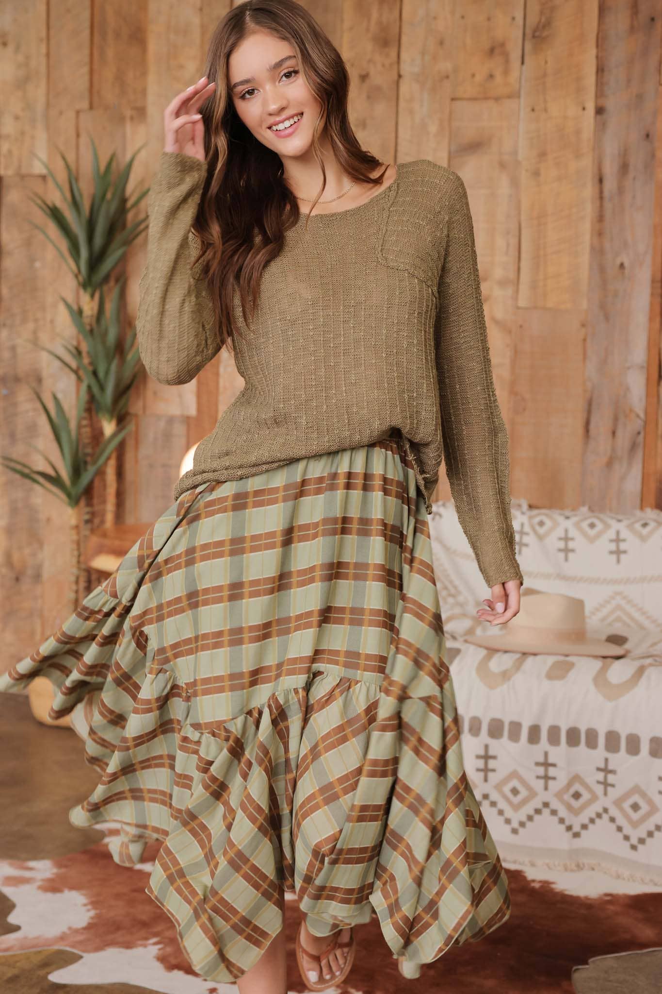 Front view of model wearing olive distressed lightweight sweater with ribbed knit texture and raw edge hem, paired with a green and brown plaid ruffle skirt and lace-up sandals.”