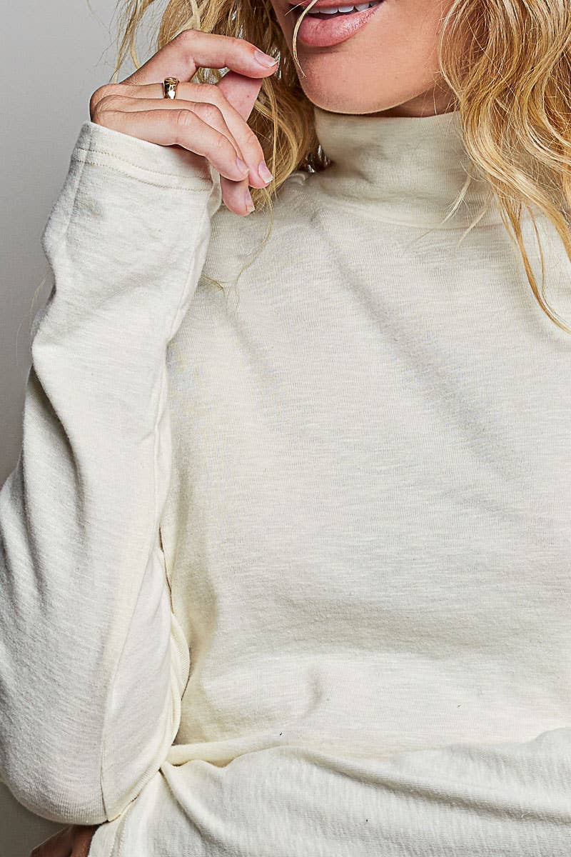 A close-up of a woman wearing a cream-colored, long-sleeve mock neck top. The image shows her hand with rings, her mouth, and her blonde hair against a neutral light gray background.