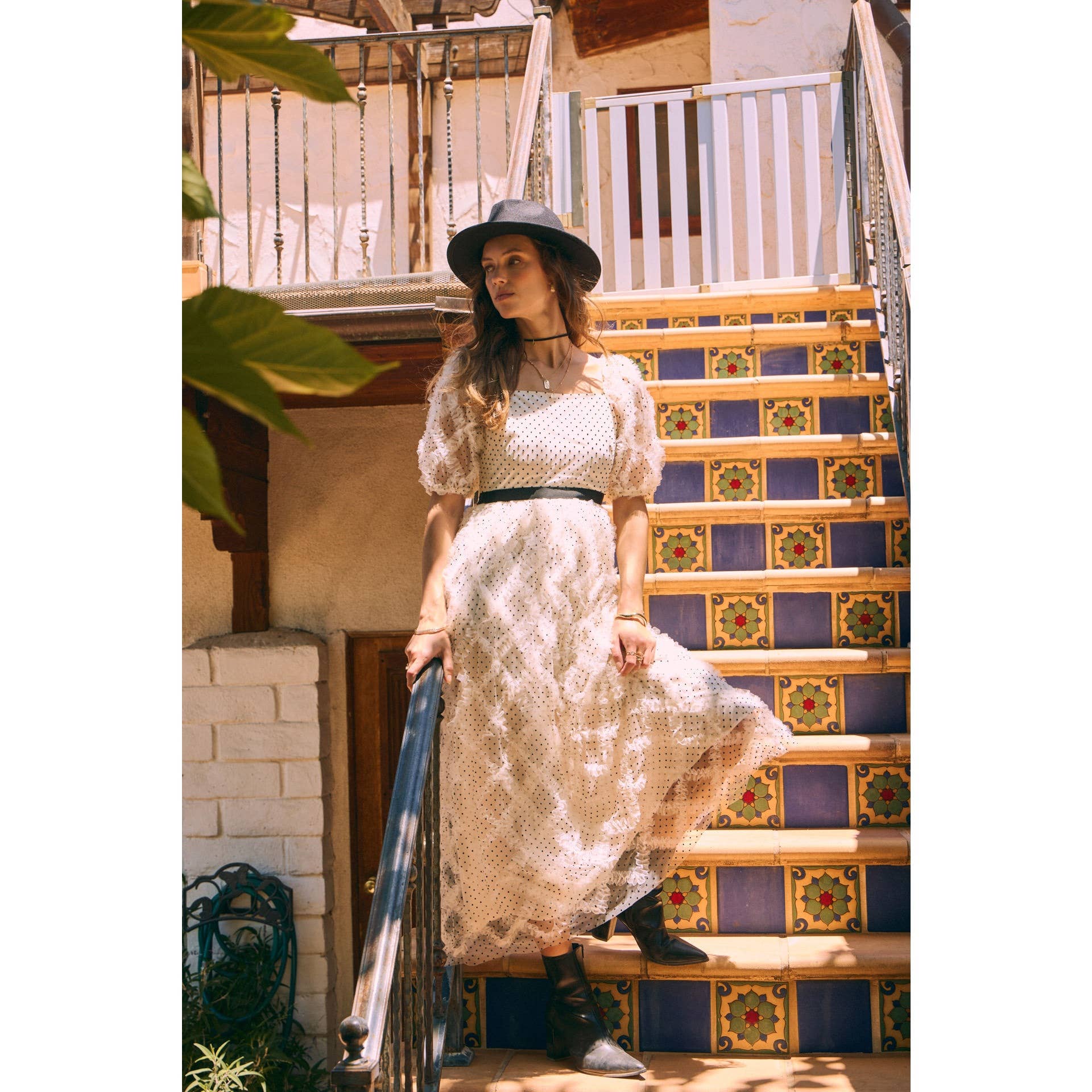 Front view — cream polka-dot tulle midi dress with puff sleeves and black belt; styled with black hat, ankle boots, pearl clutch; leaning by wooden doors.