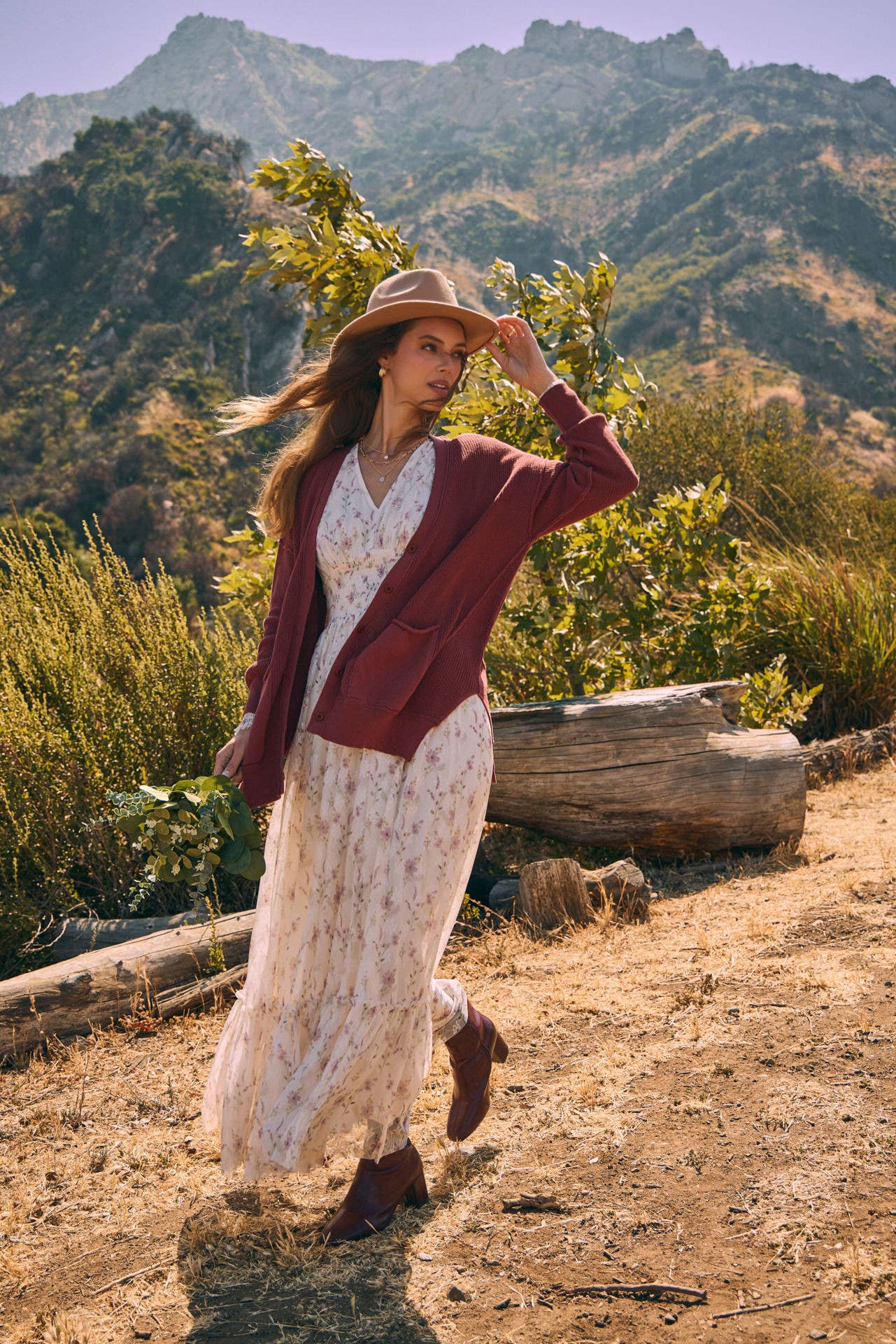 Front view — cream floral maxi dress with smocked waist and ruffle hem; mauve cardigan, tan hat, holding a greenery bouquet outdoors.