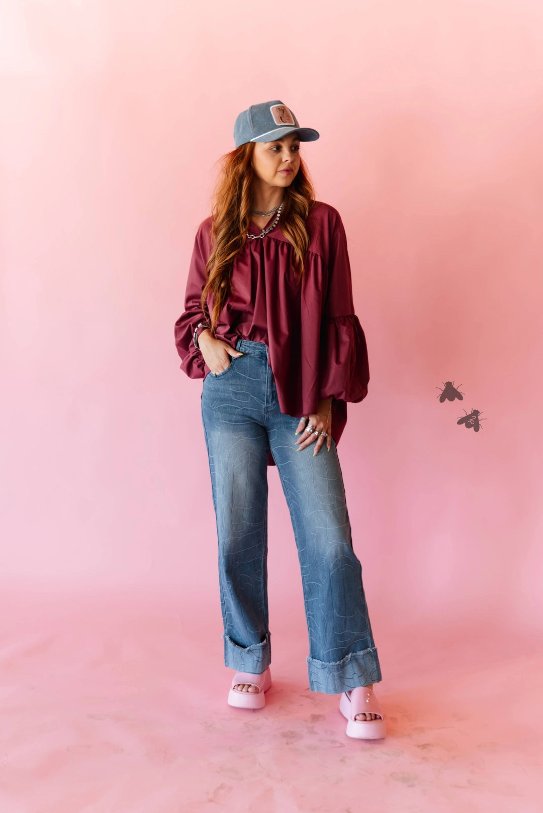 A woman stands against a pink studio backdrop wearing a burgundy long-sleeve blouse with ruffled shoulders, blue wide-leg jeans, a gray baseball cap, and pink slip-on shoes.