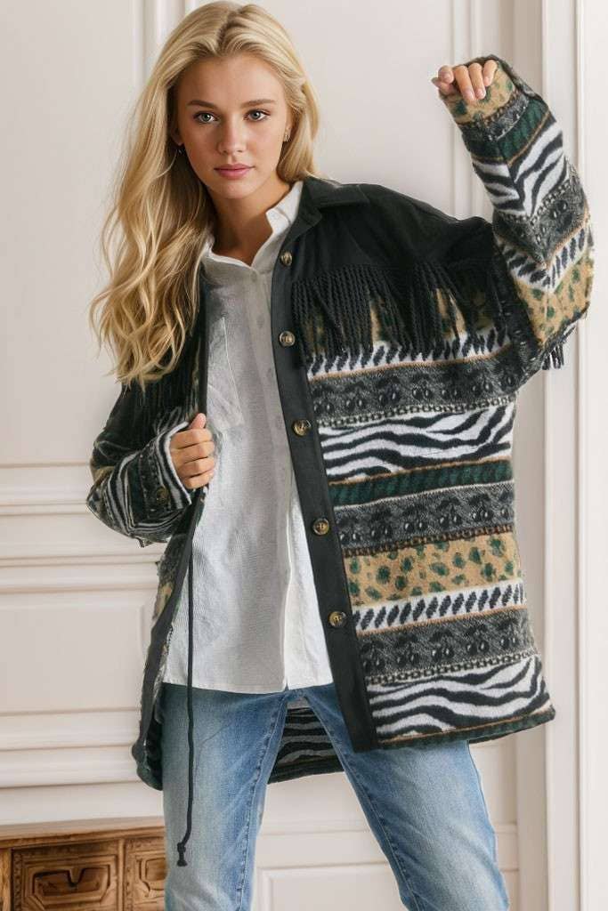 Brown Aztec print shacket with fringe detail, button closure, and patterned sleeves, styled with cream pleated pants while seated on a sofa.