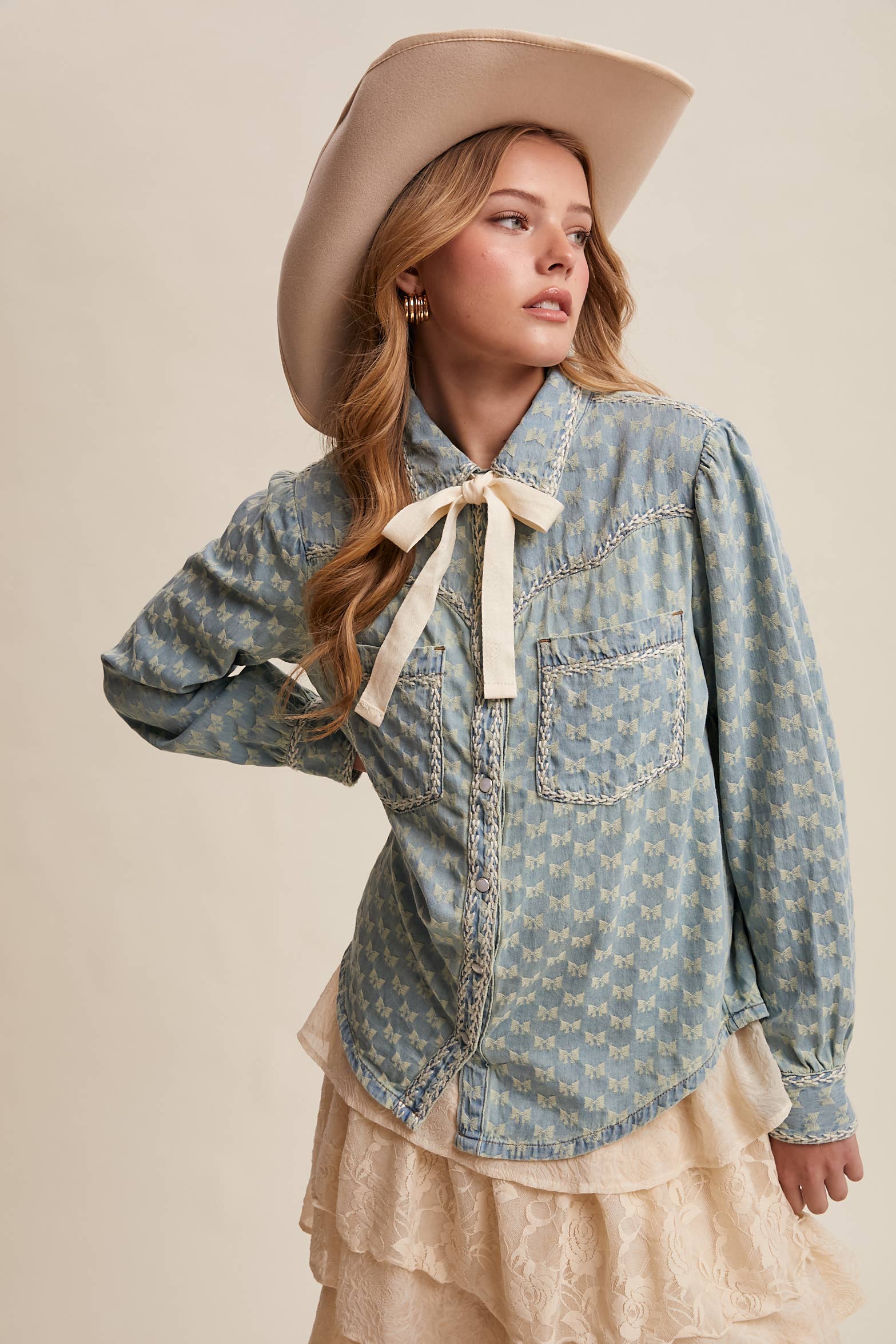 Light-wash denim blouse with cream bow-tie collar, tonal mini-bow pattern, braided contrast stitching, snap front, and two chest pockets.