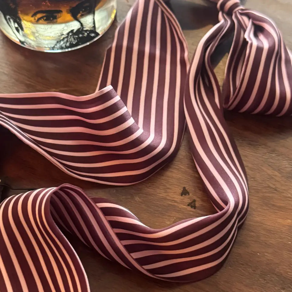 A striped necktie with alternating dark burgundy and light pink stripes, draped on a wooden tabletop next to a glass of whiskey.