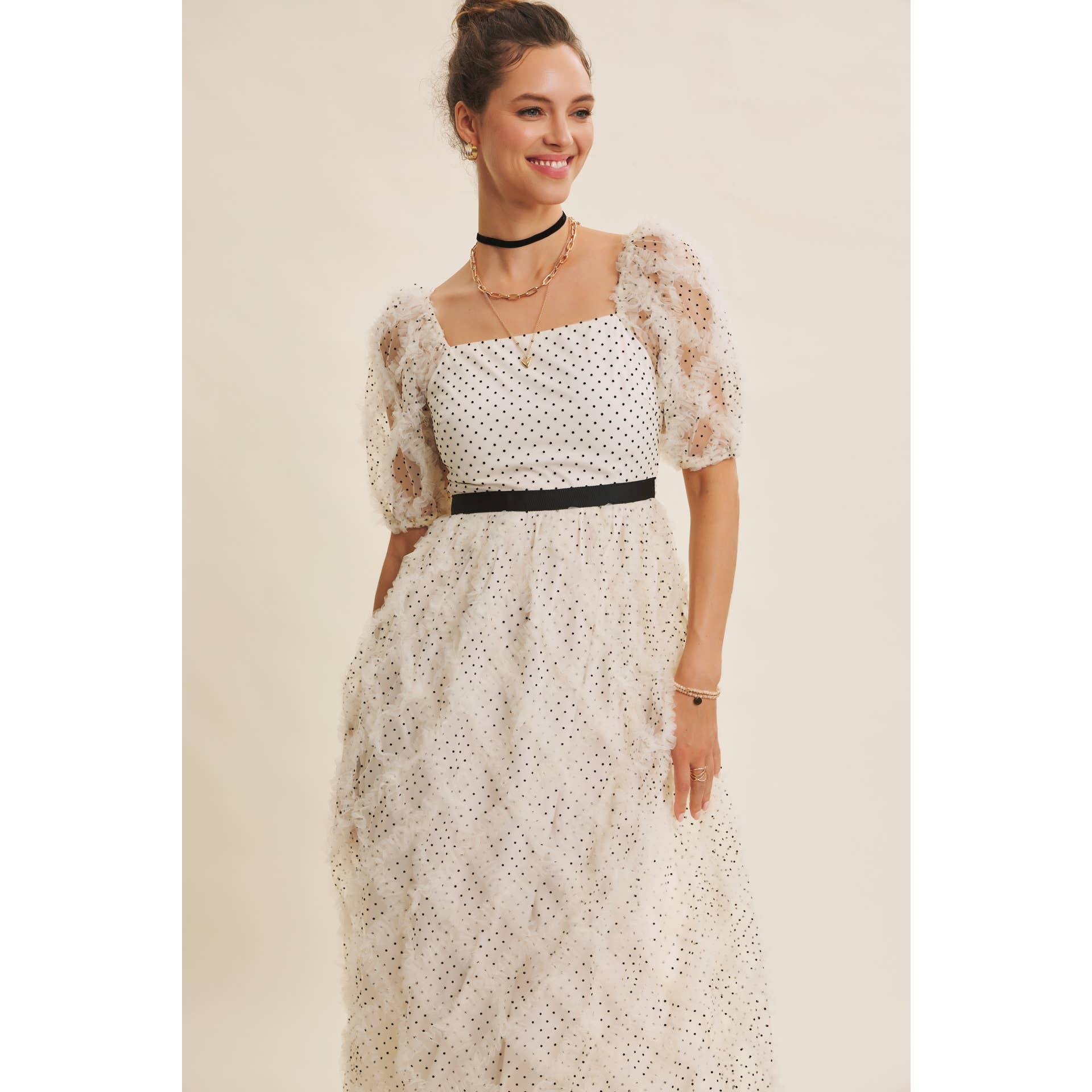 Front view — cream polka-dot tulle midi dress with puff sleeves and black belt; styled with black hat, ankle boots, pearl clutch; leaning by wooden doors.