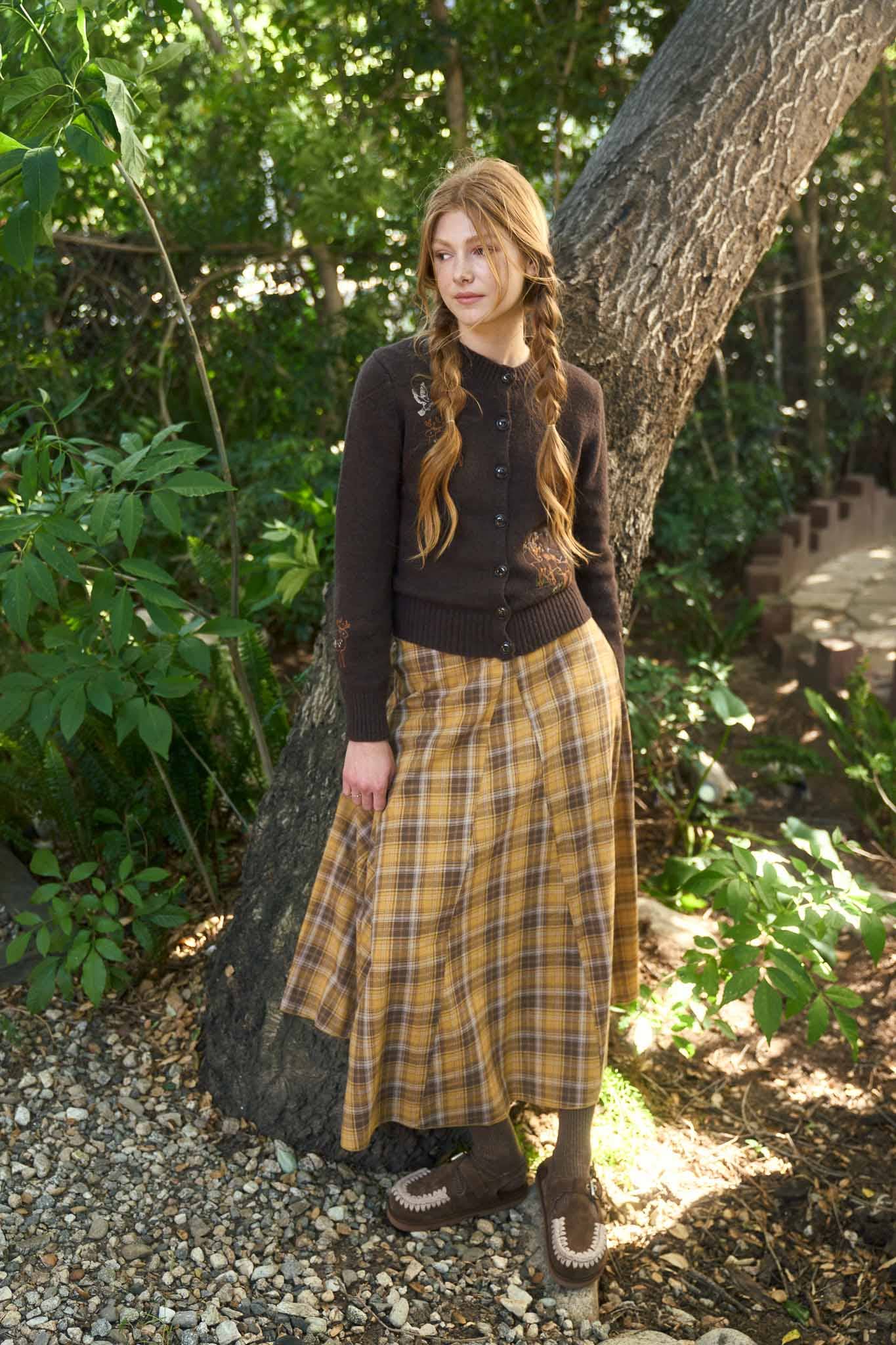 Woman in a brown cardigan and plaid skirt standing in a forested area.