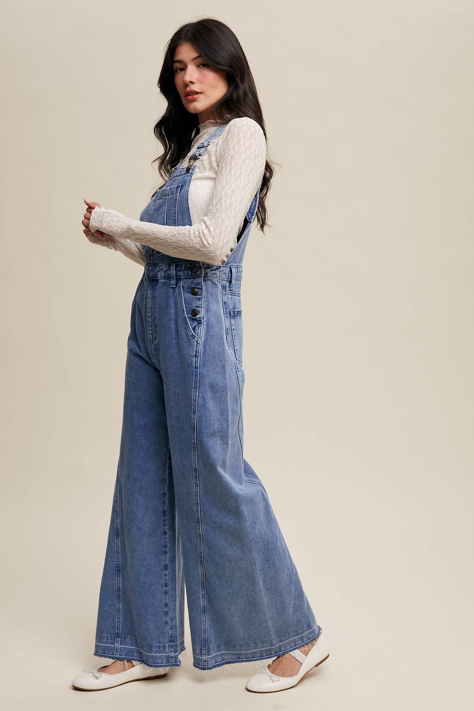 Side view- Light-wash wide-leg denim overalls with adjustable straps, front bib pocket, side buttons, and slant pockets, finished with a raw hem and layered over a cream long-sleeve top.