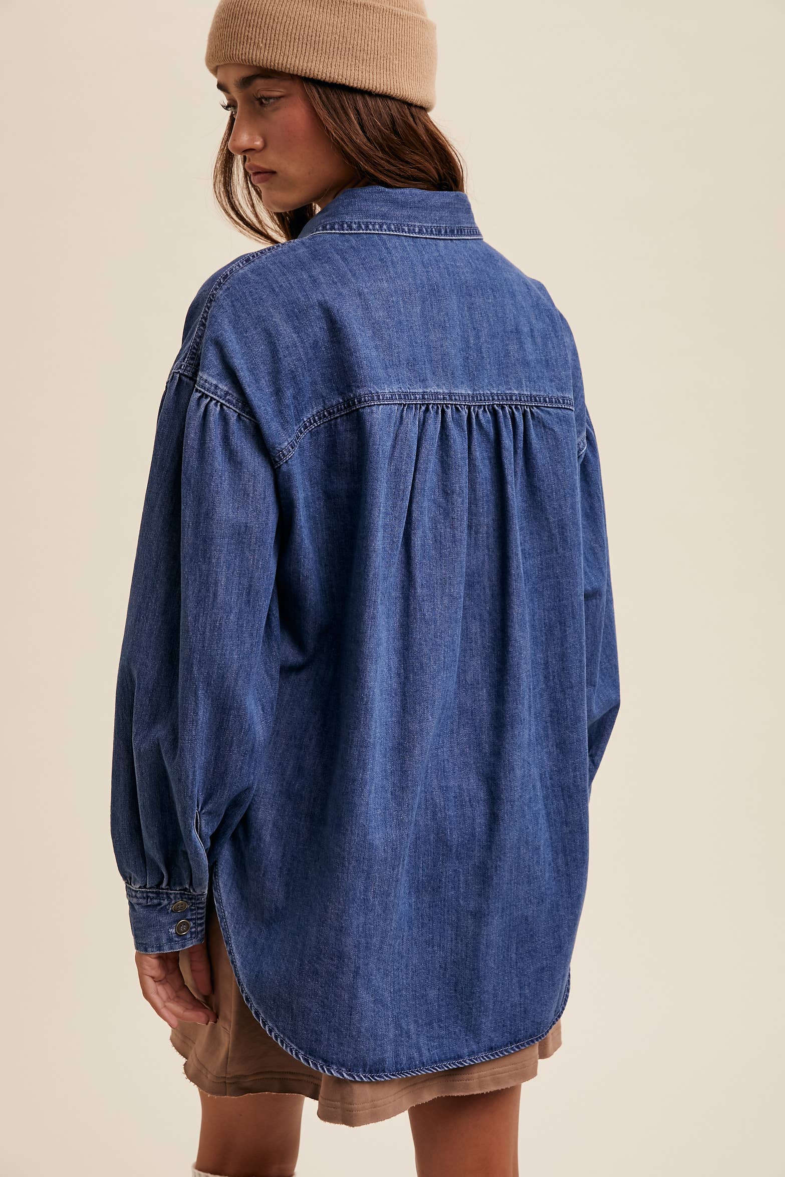 Back view- Medium-wash oversized denim shacket with button front, drop shoulders, balloon sleeves, side pockets, and a curved shirttail hem with side slits.