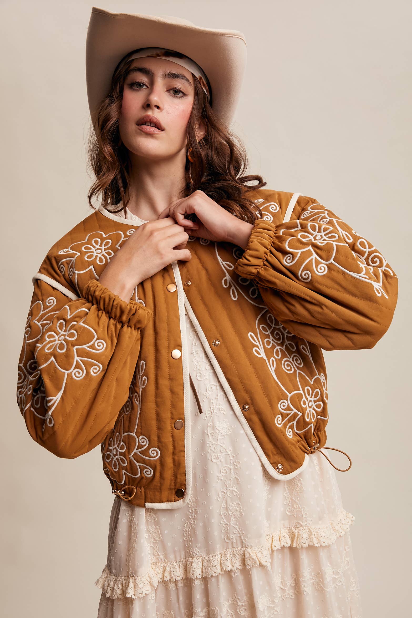 Boho caramel-brown jacket with cream floral swirl embroidery, ivory piping, and snap buttons, styled over a lace tie-front top with a scarf and felt hat outdoors.