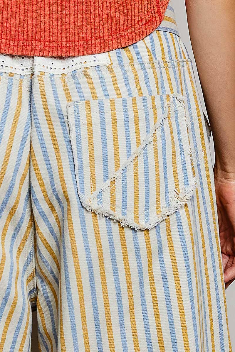 Close-up of striped pants with a frayed triangular pocket detail, worn with an orange ribbed top. The pants feature vertical stripes in light blue, mustard yellow, and off-white.