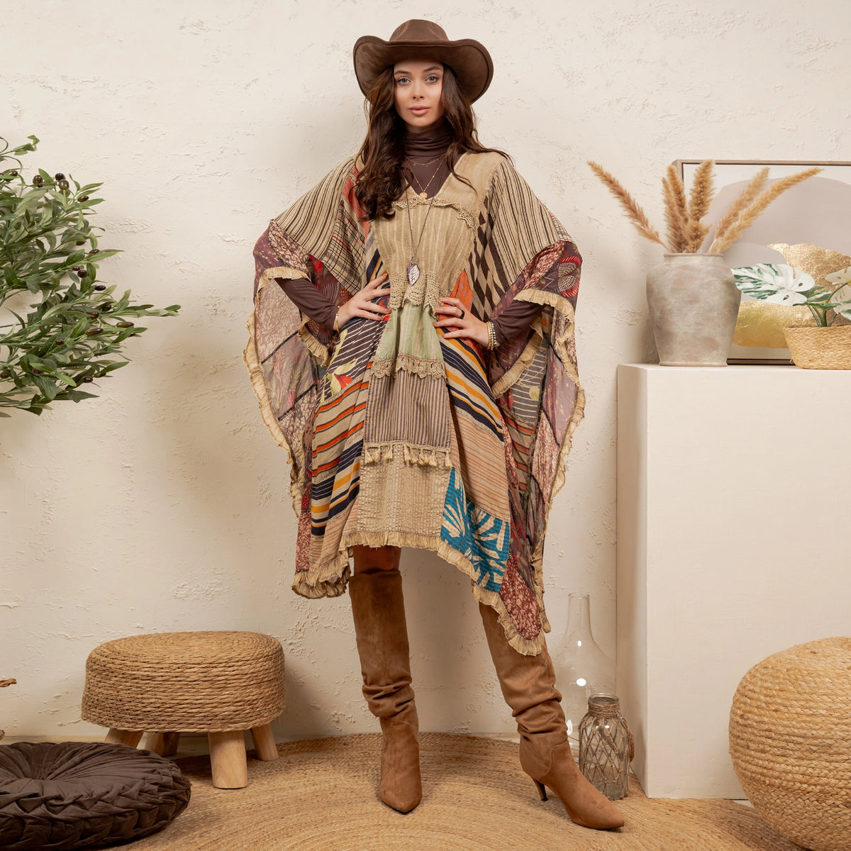 A woman stands indoors wearing a bohemian-style patchwork poncho with fringe, a brown wide-brim hat, a dark turtleneck, brown slouchy boots, and a brown hat. The room has a textured cream wall, woven stools, plants, and decorative vases.