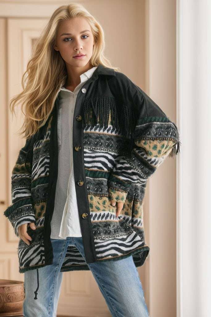 Brown Aztec print shacket with fringe detail, button closure, and patterned sleeves, styled with cream pleated pants while seated on a sofa.