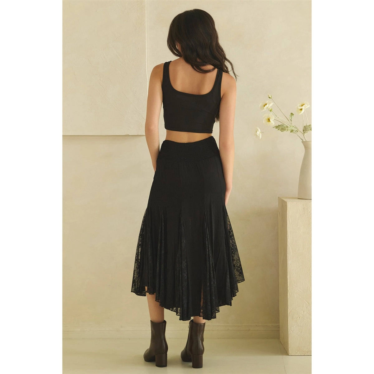 Calla Lace Godet Maxi Skirt in Black by Oli & Hali — back view showing structured waistband, flaring black satin and floral lace godet panels, and asymmetric handkerchief hem with ankle boots