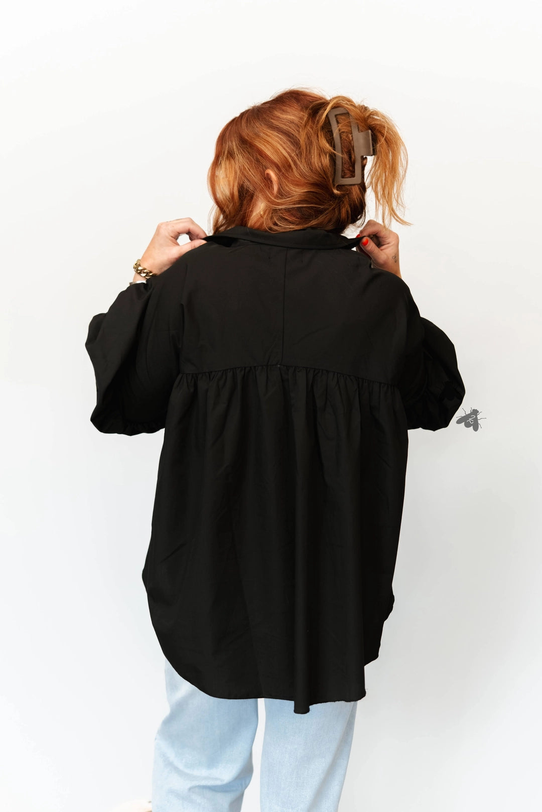 A person viewed from behind wearing an oversized black long-sleeve blouse with a gathered waist seam and light blue jeans, standing against a plain white background.