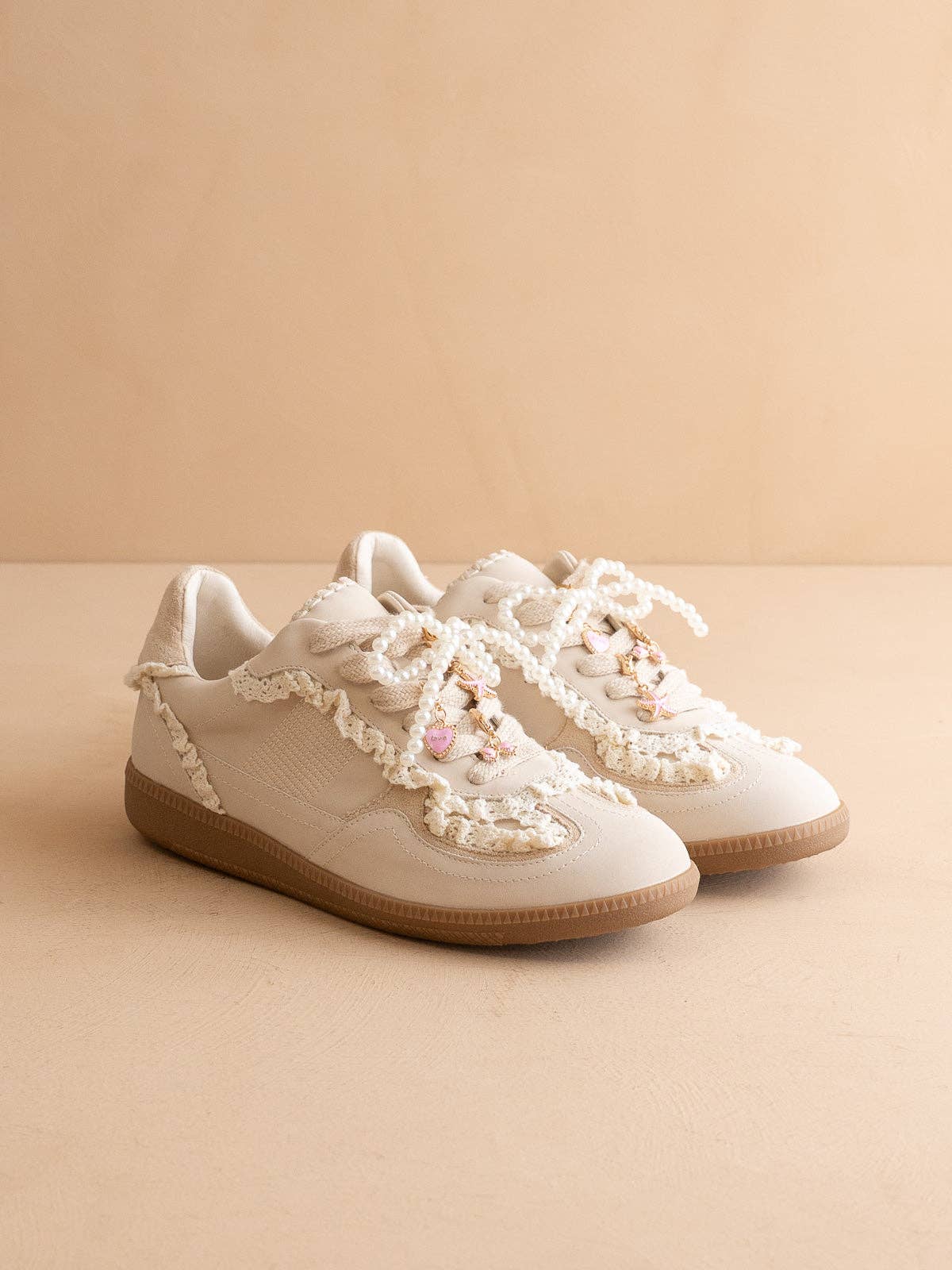 Pair of ivory lace-trim low-top sneakers with pearl-beaded laces, pink heart charms, and gum soles on a neutral backdrop.